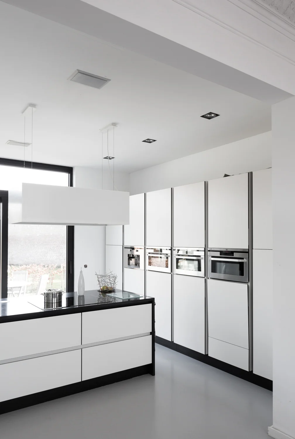 Modern minimalist kitchen with white cabinetry, black countertops, built-in ovens, and a large window showing an outdoor area.