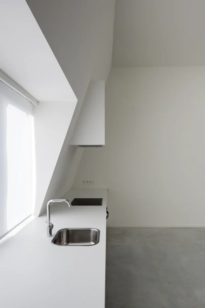 Minimalist kitchen with white countertops, a stainless steel sink, and a white window blind, featuring an angled ceiling design.