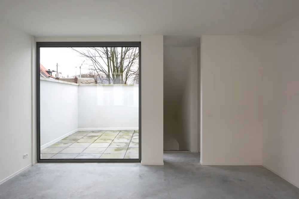 Empty room with large glass sliding door leading to a small outdoor patio with tiled flooring and a white fence, in a modern apartment or house.