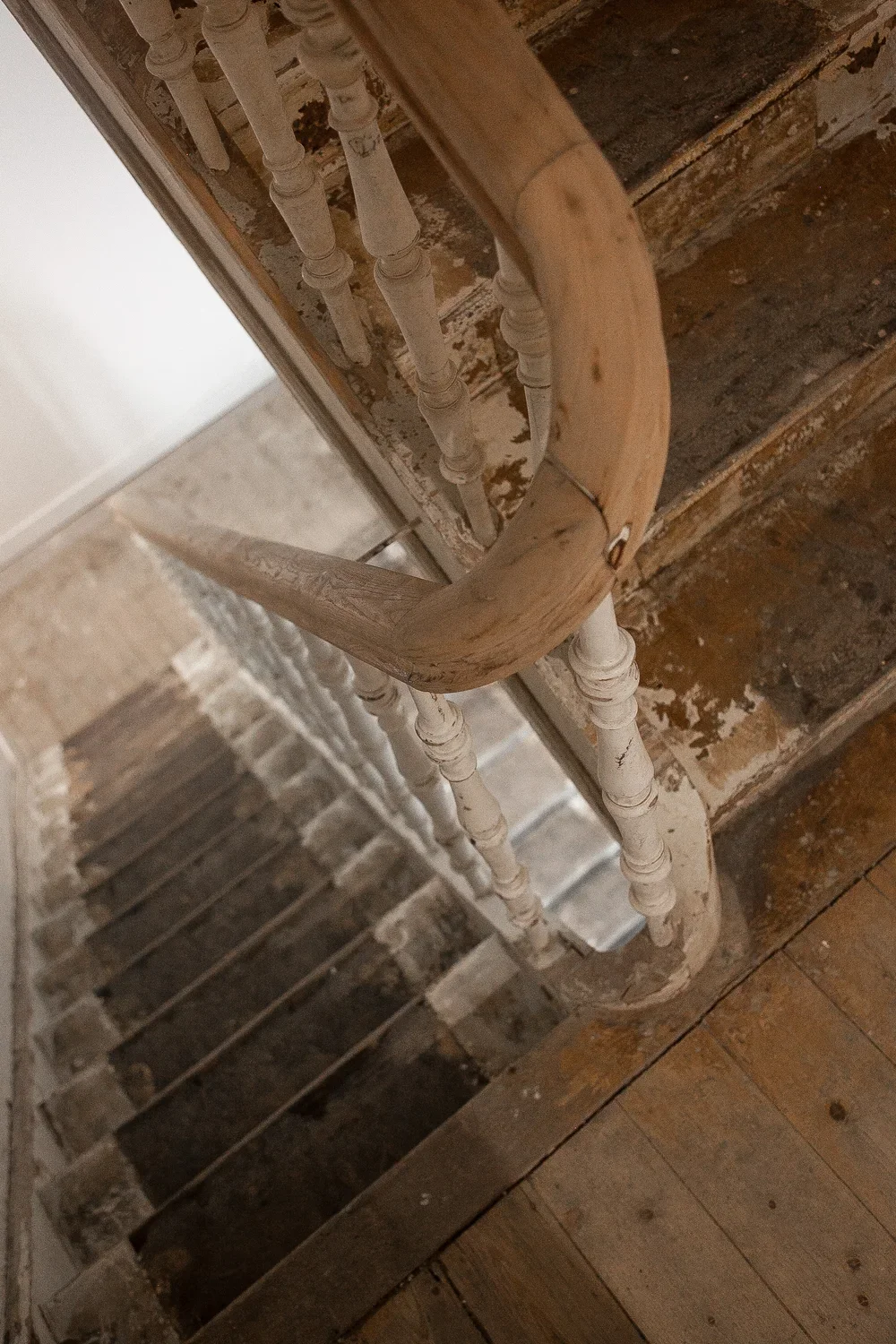 An old wooden staircase with a curved banister, viewed from above, with exposed brick wall and peeling paint.
