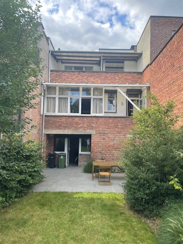 View of a multi-story brick apartment building with a backyard patio area, plants, and a bench, under a partly cloudy sky.