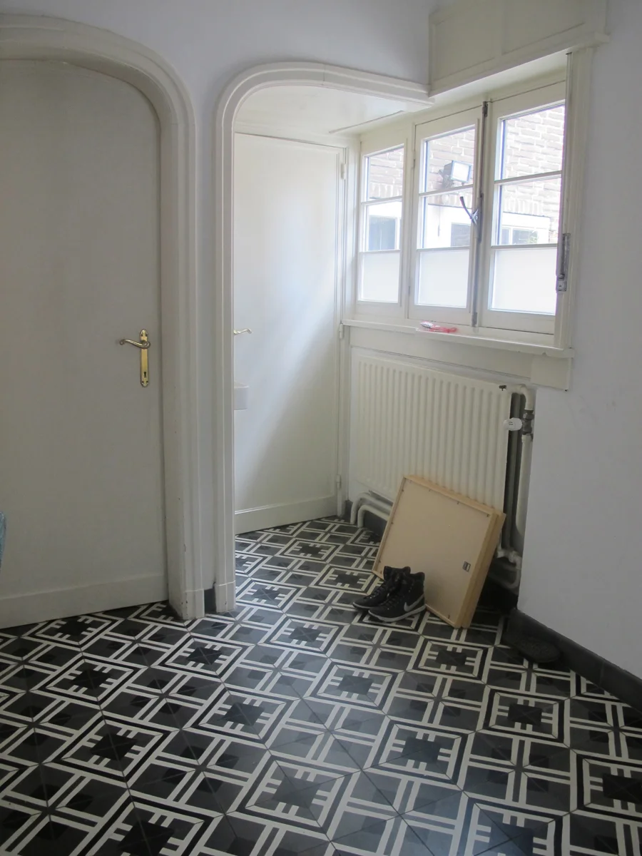 A small room with a patterned black and white tiled floor, a white door, and a window letting in natural light. There is a pair of black boots and a wooden frame or panel leaning against the wall near the window. The room has white walls and a radiator beneath the window.