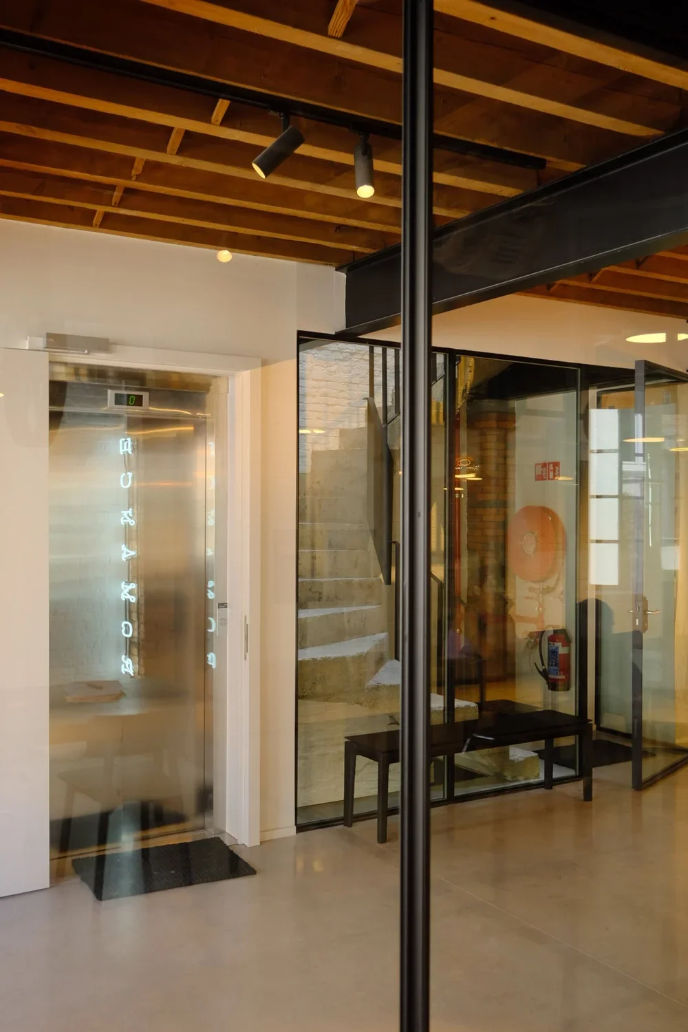 Modern building interior design featuring an integrated elevator, glass partition walls, exposed wooden ceiling beams, and contemporary stairs, by Tom Naeyaert Architecture.
