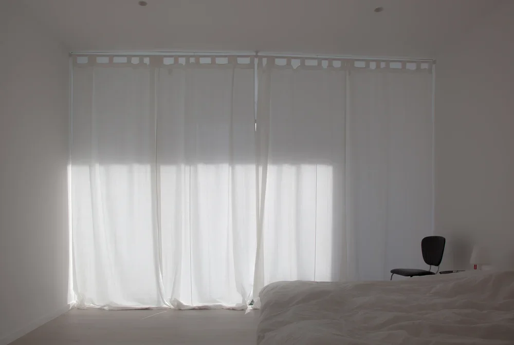 A bedroom with white curtains, a bed, and a black chair.