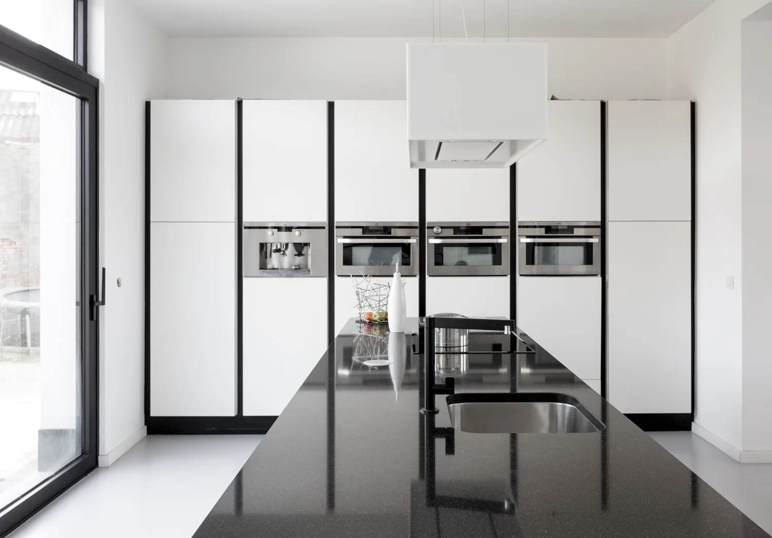 Modern kitchen with black island countertop, stainless steel sink, white cabinets, and built-in ovens, with a sliding glass door on the left.