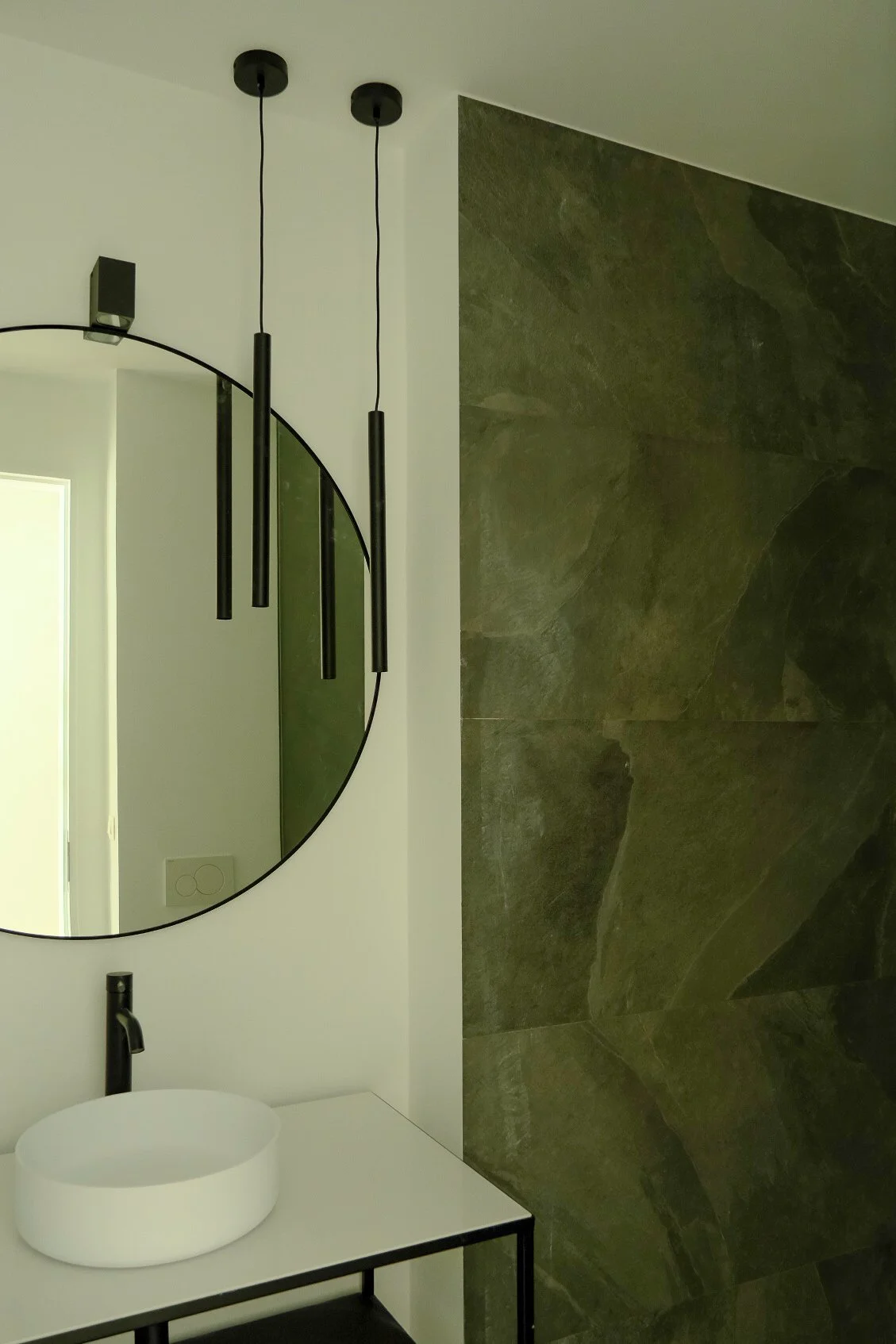 Modern bathroom interior design featuring a round mirror, black lighting fixtures, and a dark green stone wall, by Tom Naeyaert Architecture.