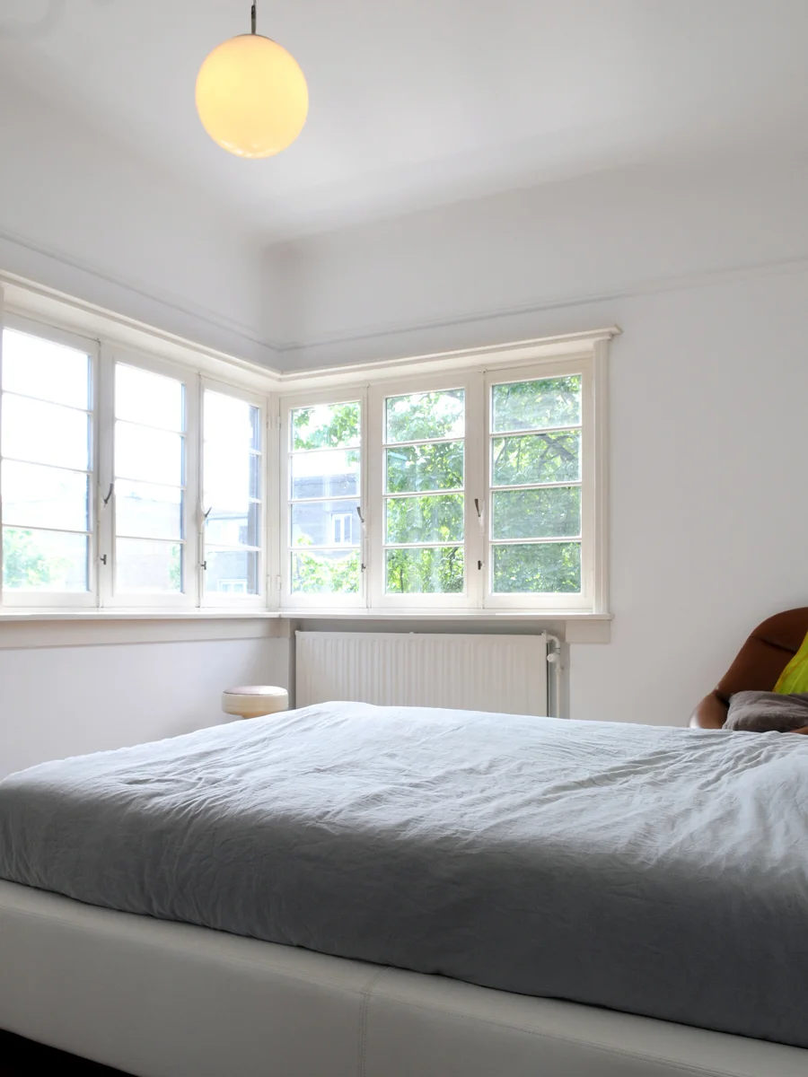 A bright bedroom with large windows, a bed with gray bedding, a small round stool, and a brown chair with pillows, illuminated by a ceiling light