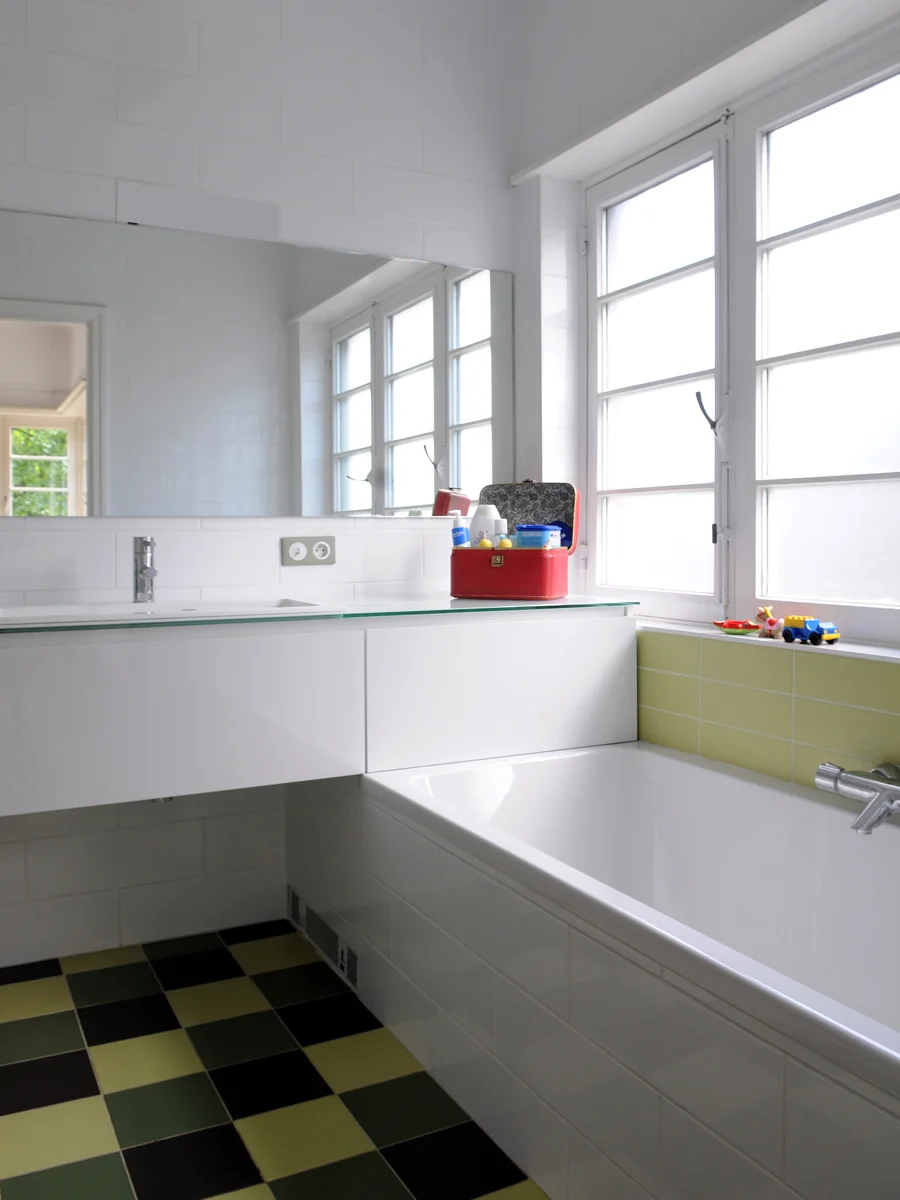 A modern bathroom with a bathtub, white walls, and large windows letting in natural light. There are small toys on the windowsill and a red container with toiletries on the counter.