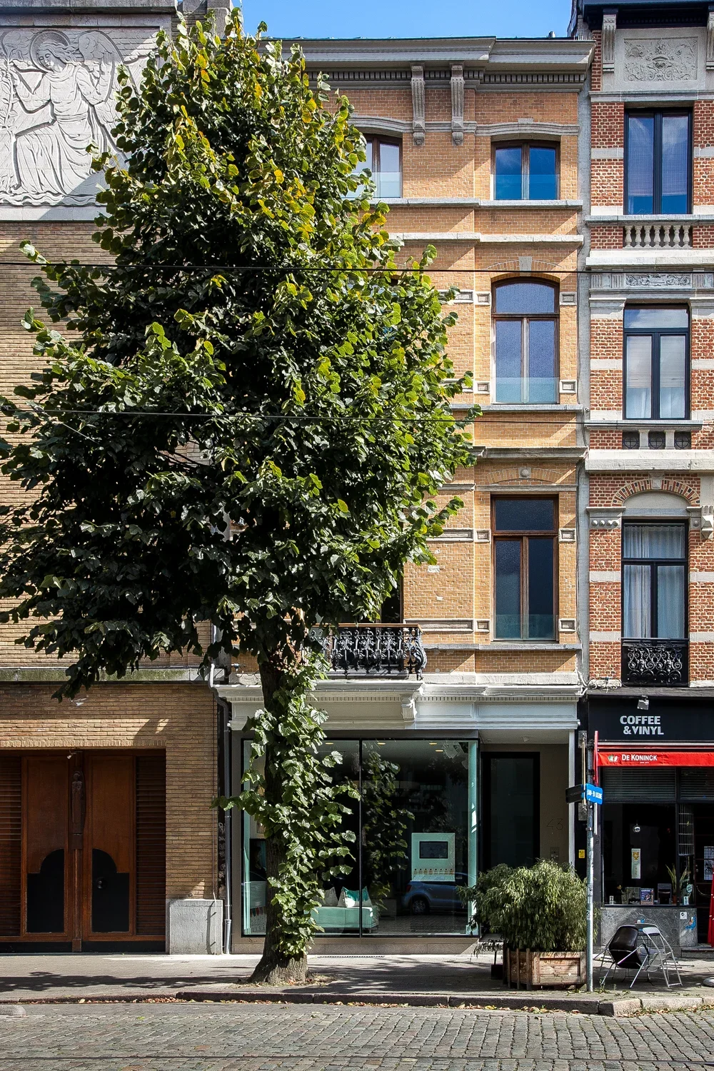 Street view of a multi-story brick building with large windows and ornate architectural details, partially obscured by a leafy tree, with a café and a plant on the sidewalk.