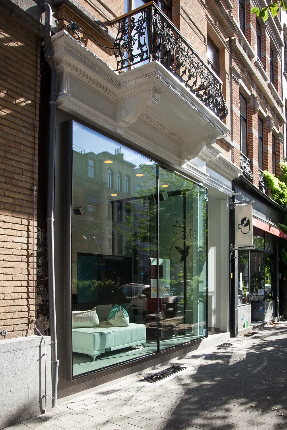 City street scene with a storefront featuring large glass windows, white decorative molding, and a balcony with ornate black wrought iron railing above. The real estate reflects surrounding buildings and a sidewalk shaded by trees.