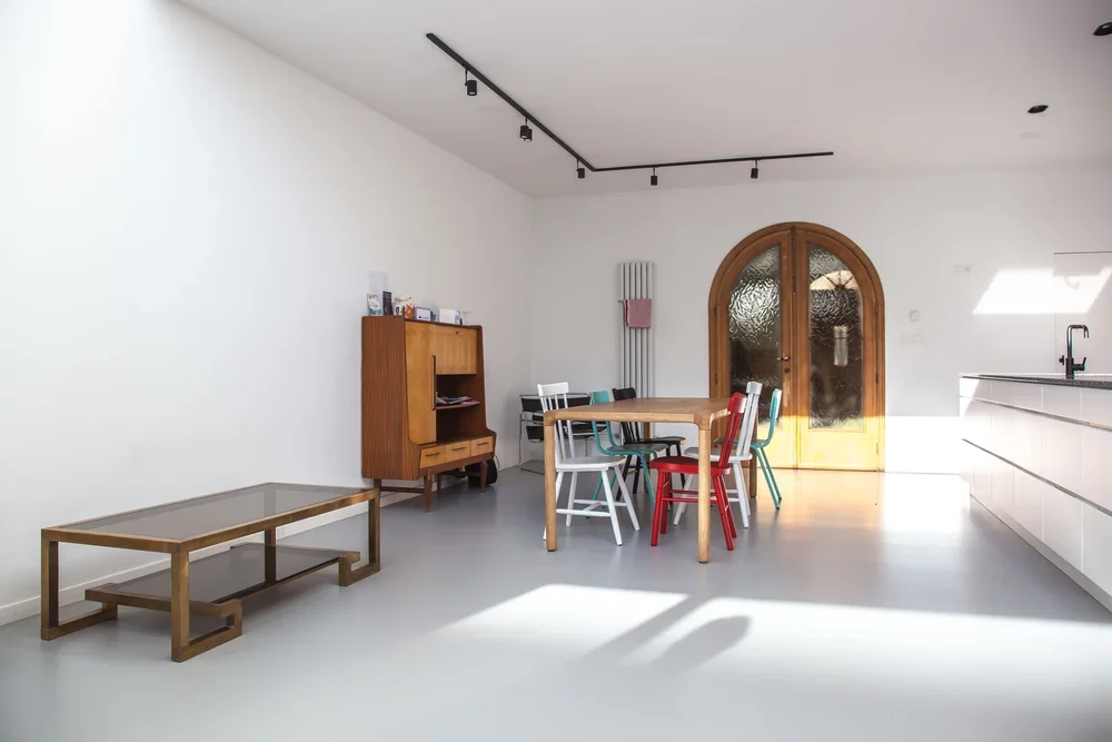 Minimalist dining area with a round wooden table, colorful chairs, a wooden cabinet, a glass-top coffee table, and a white kitchen counter with a sink, arched wooden door, and natural light.