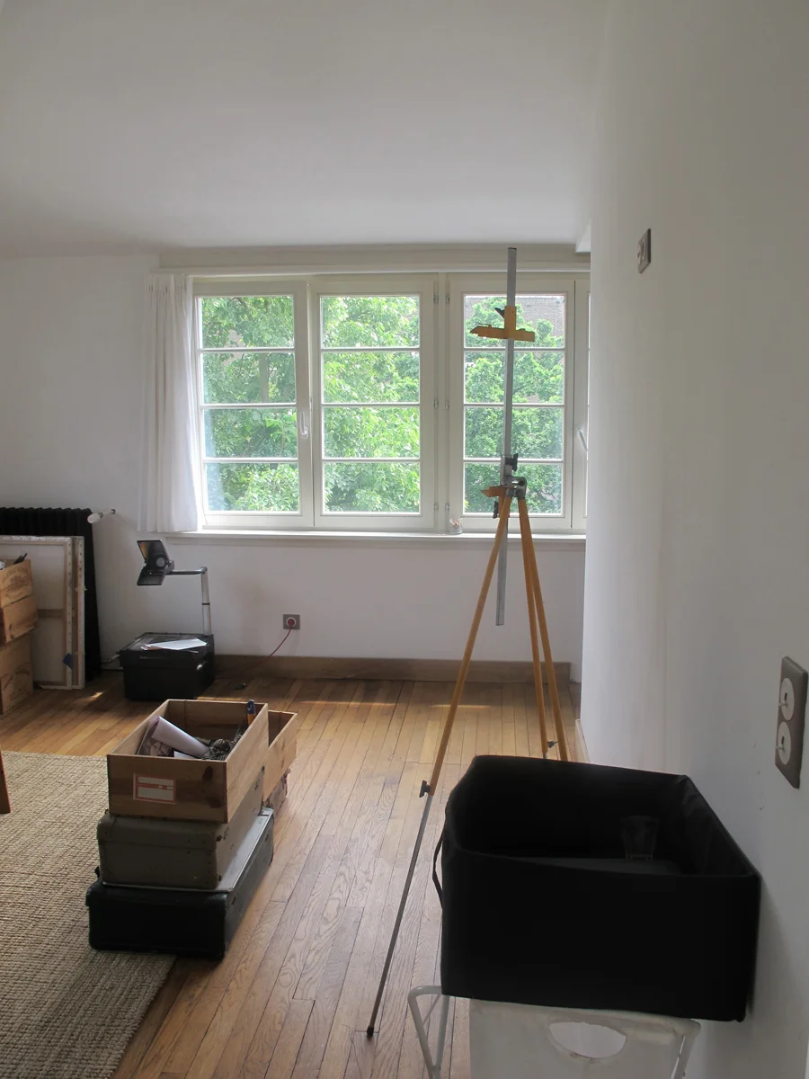 A room with large window overlooking green trees, with a tripod and measuring instrument set up inside, and various boxes and objects on the wooden floor.