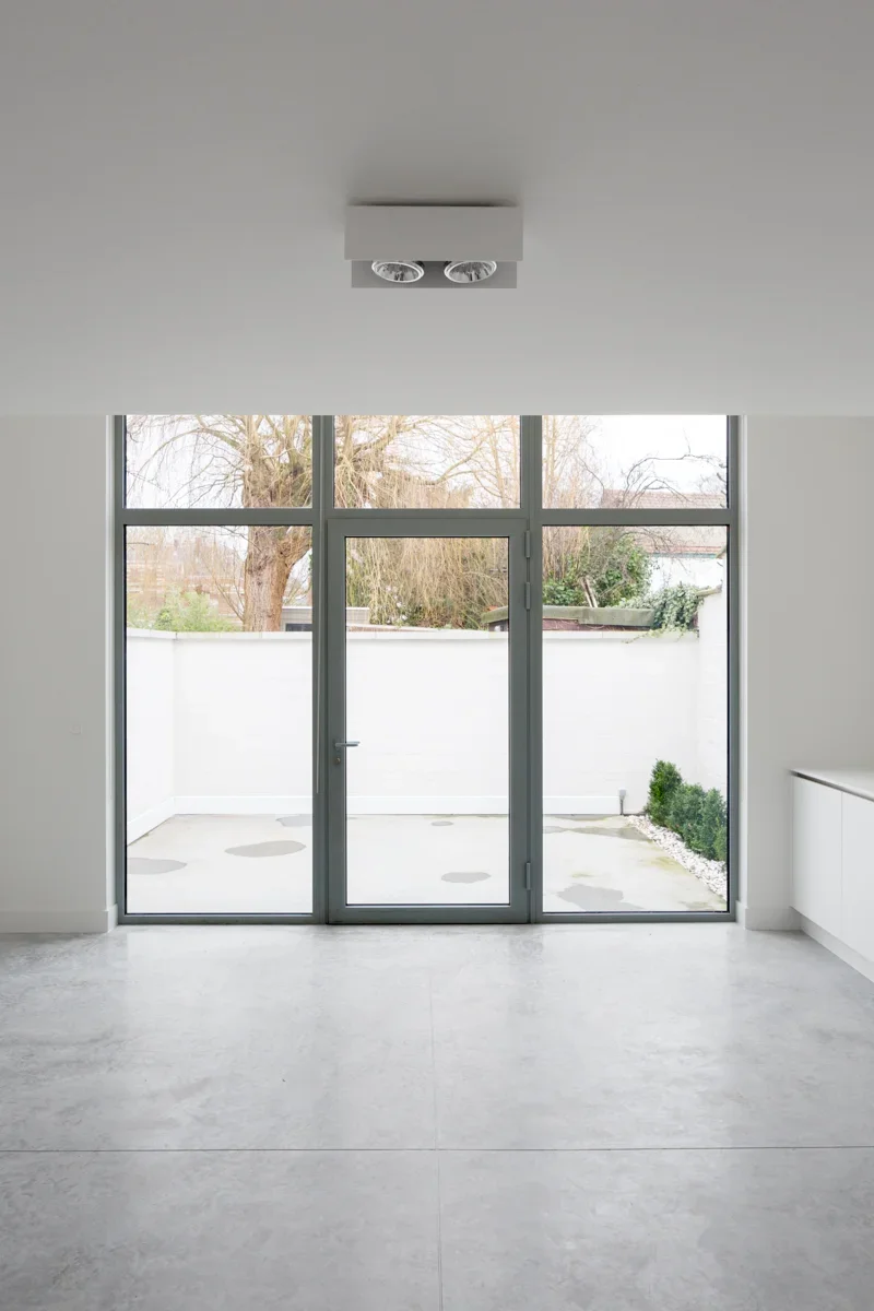 Empty modern room with large glass sliding door leading to outdoor patio, white walls, and light gray tiled floor.