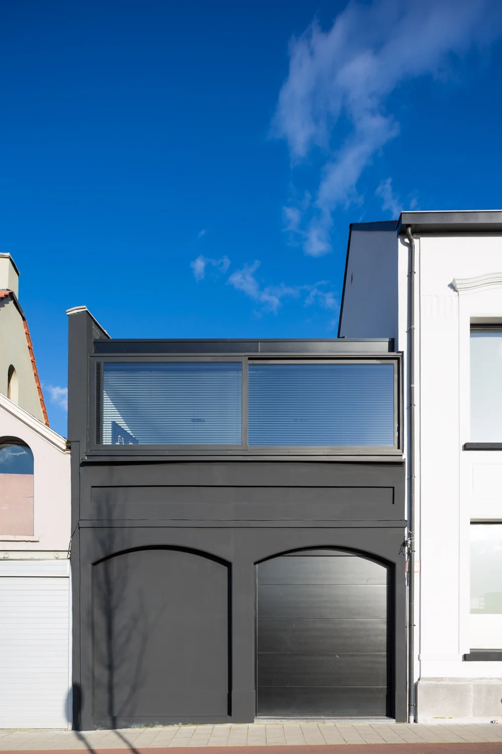 Modern building with black facade and large windows, situated between older buildings under a bright blue sky with a few clouds.