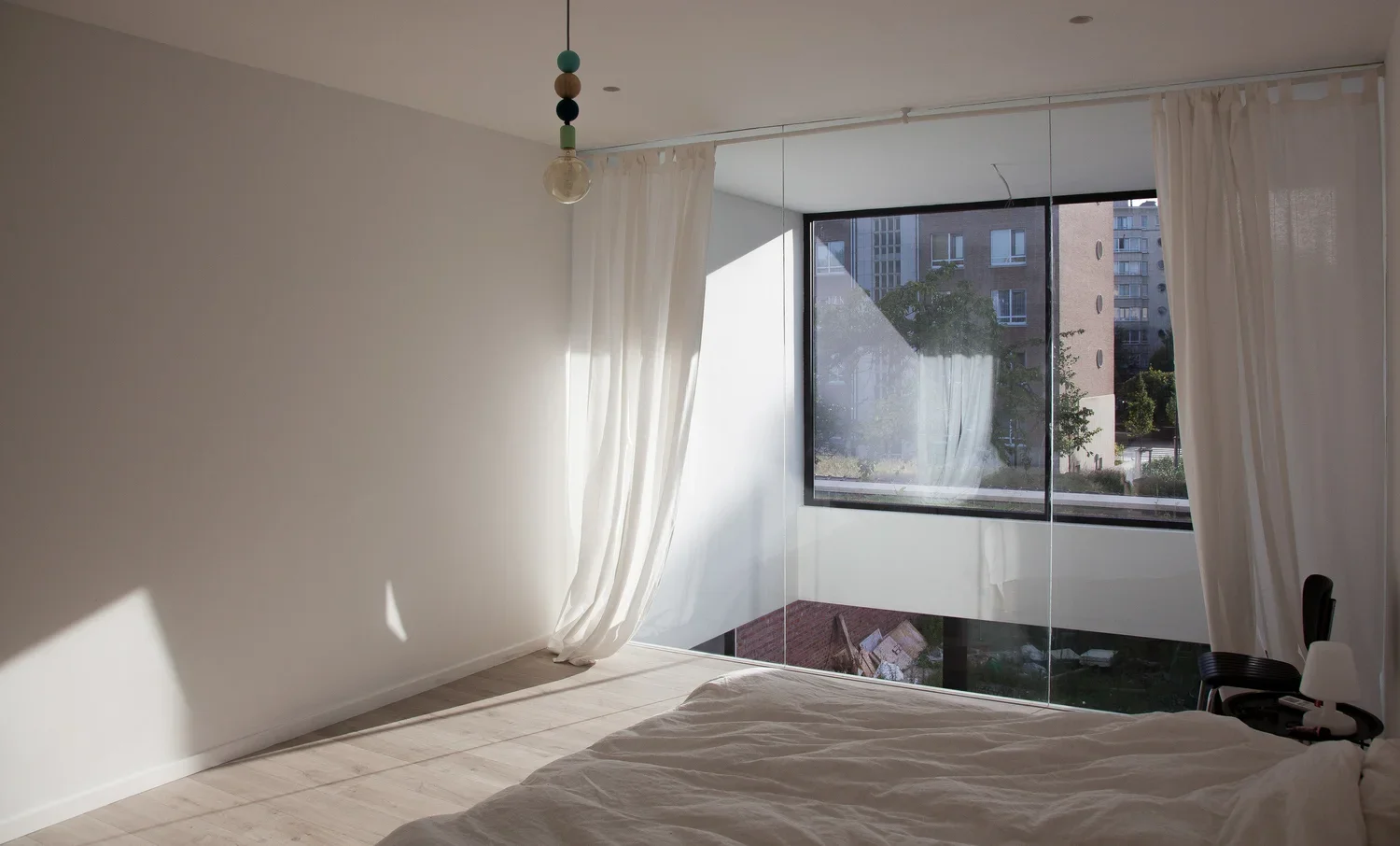 A minimalist bedroom with white walls, a large window with white curtains, and a bed with white bedding. Sunlight streams through the window, casting shadows on the floor.