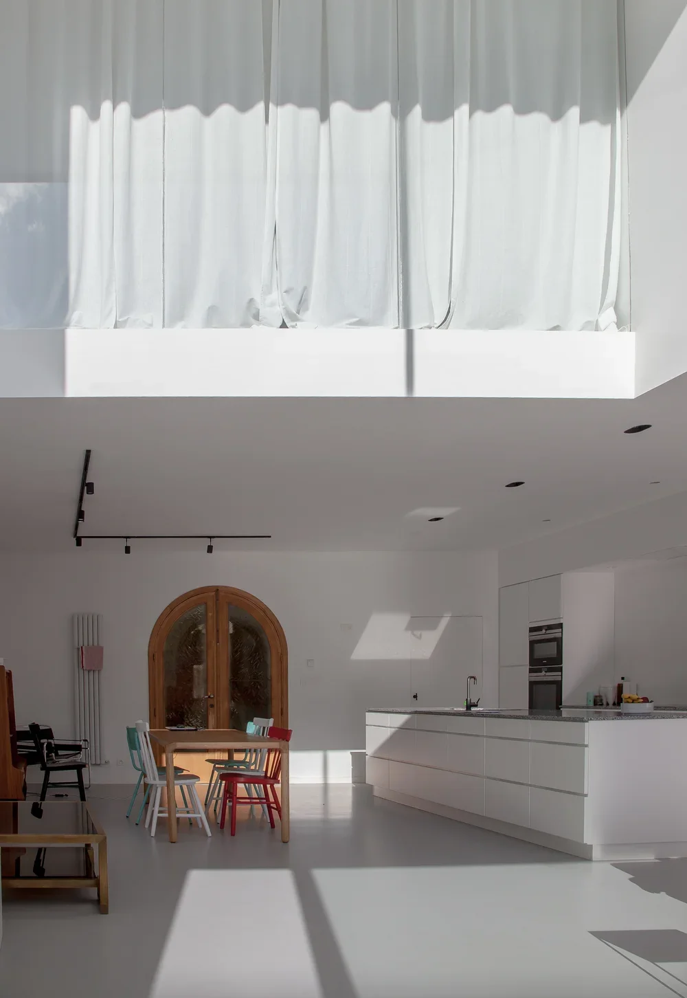 Modern, minimalist kitchen and dining area with white walls, a wooden arched door, a white kitchen island, and colorful chairs around a wooden table, sunlight casting shadows.