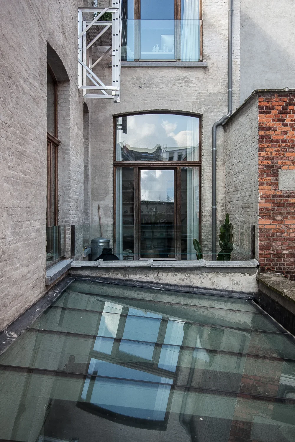 Exterior view of an urban building with large windows, glass balcony, brick and concrete walls, a rooftop garden with cactus plants, and a sloped glass roof reflecting the sky.