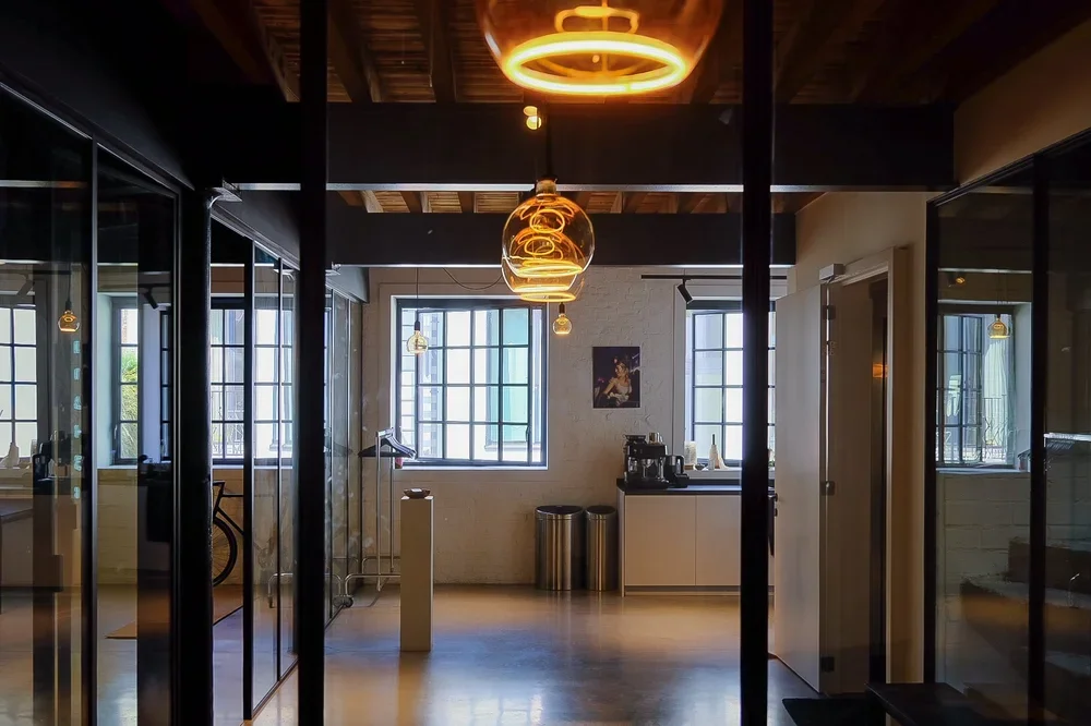 Contemporary workspace design featuring glass doors, minimalist pendant lights, and large windows maximizing natural light, by Tom Naeyaert Architecture.