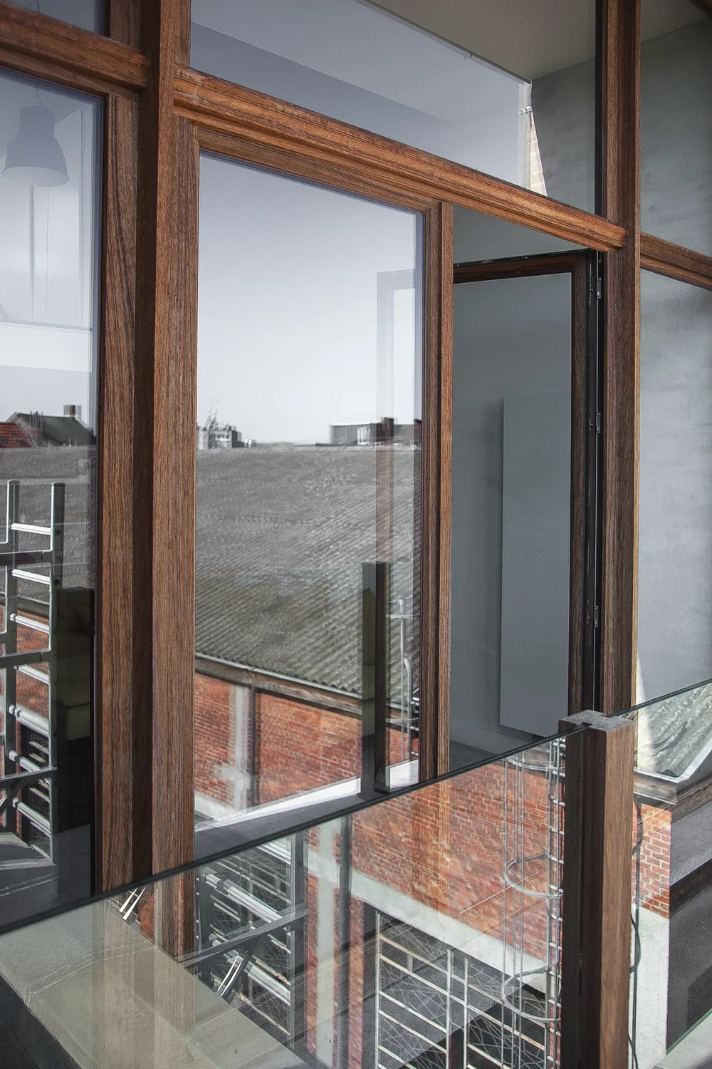 Interior view of a modern building with large glass windows and wooden frames, showing a rooftop and neighboring buildings outside.