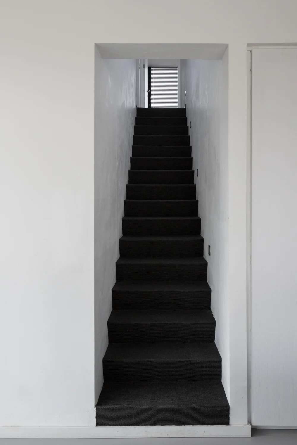 Interior view of a narrow staircase with black carpet, leading up to a window with blinds in a white wall.