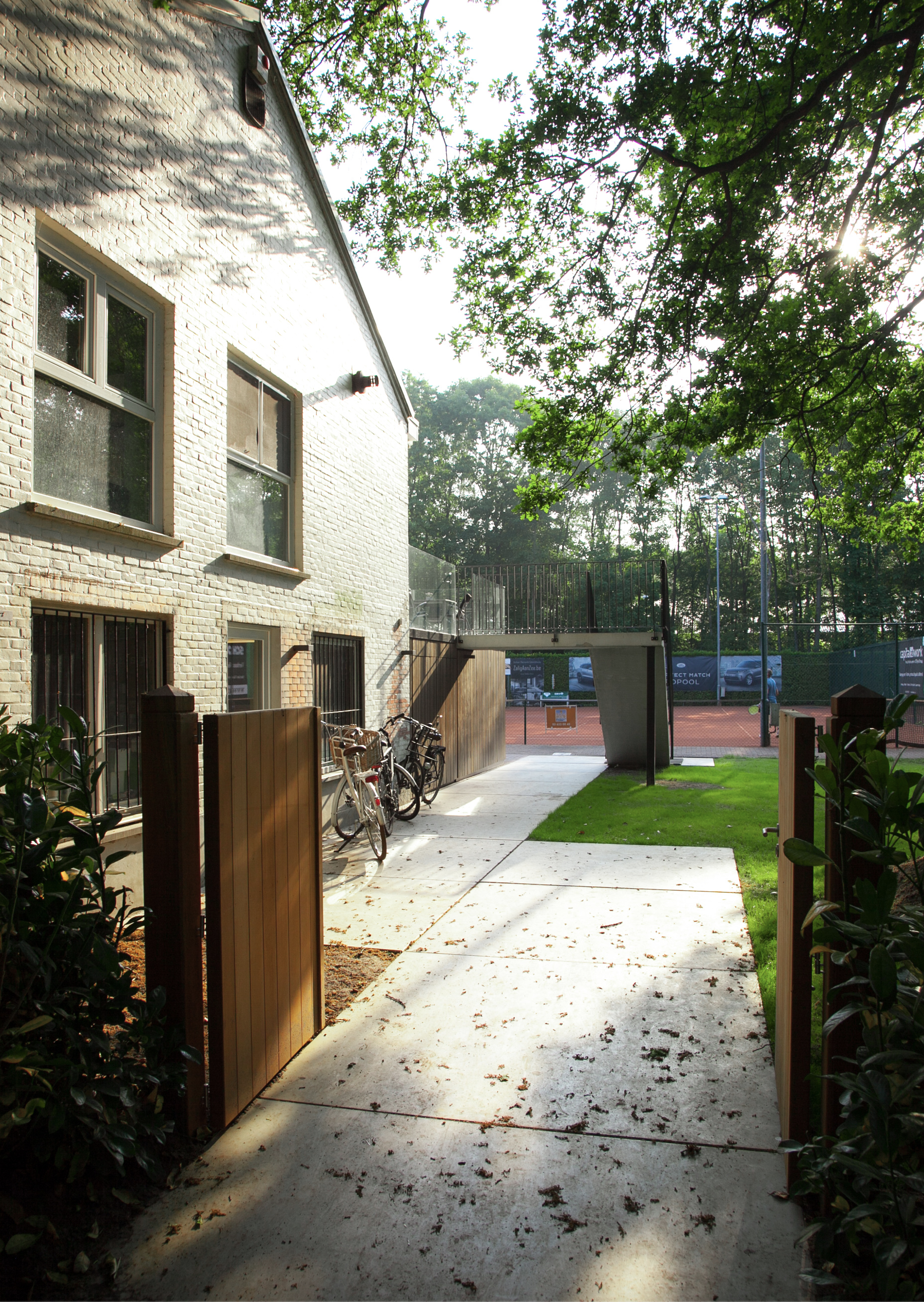 Architectural landscaping and walkway design connecting a brick building to sports facilities, by Tom Naeyaert Architecture.