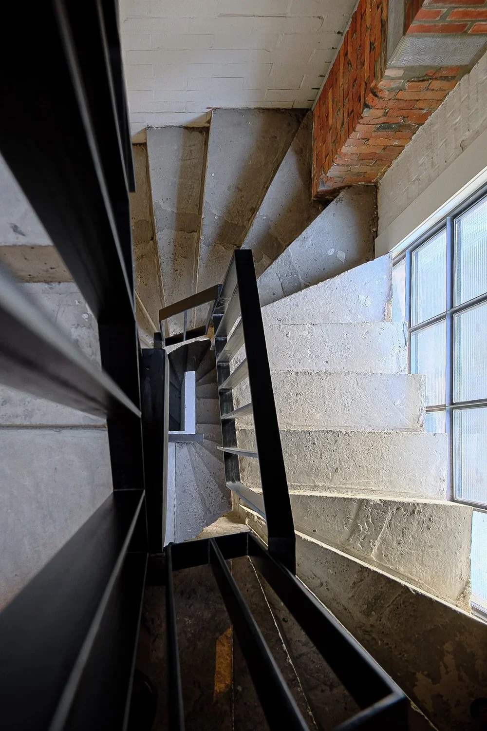 Architectural perspective looking down a modern spiral staircase with concrete steps, a brick wall corner, and a frosted glass window, by Tom Naeyaert Architecture.