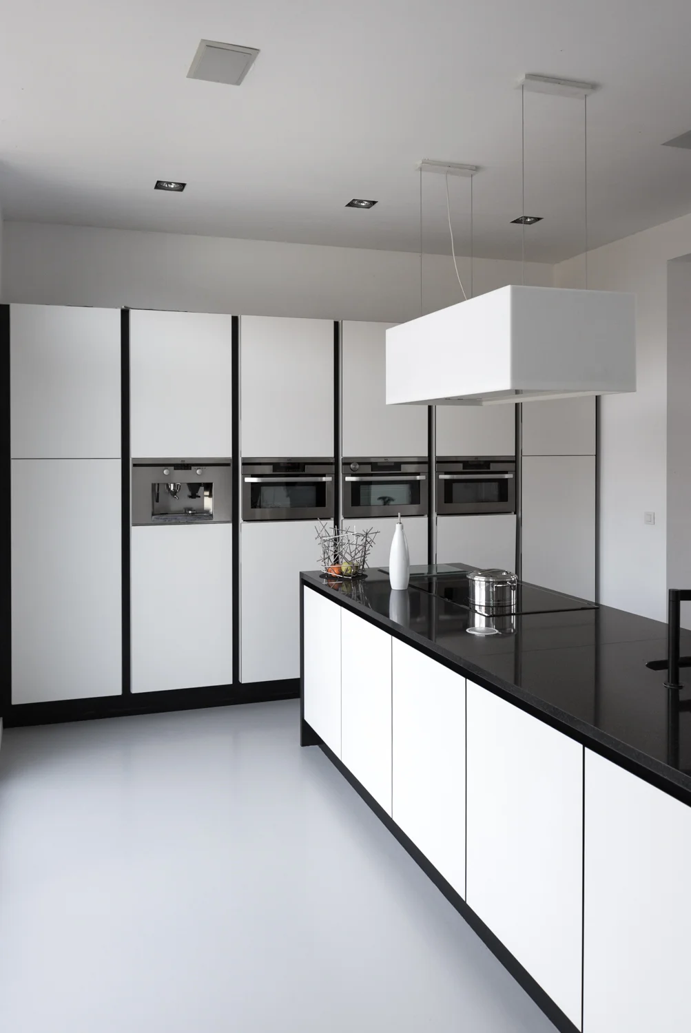 Modern kitchen with white cabinets outlined in black, black countertops, built-in oven and microwave, and a black island with decorative items and a hanging white light fixture.