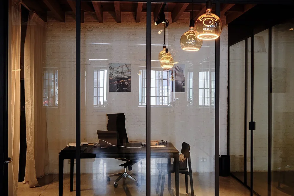 Modern office interior with corporate glass partition walls, a sleek black desk, and industrial pendant lighting, by Tom Naeyaert Architecture.