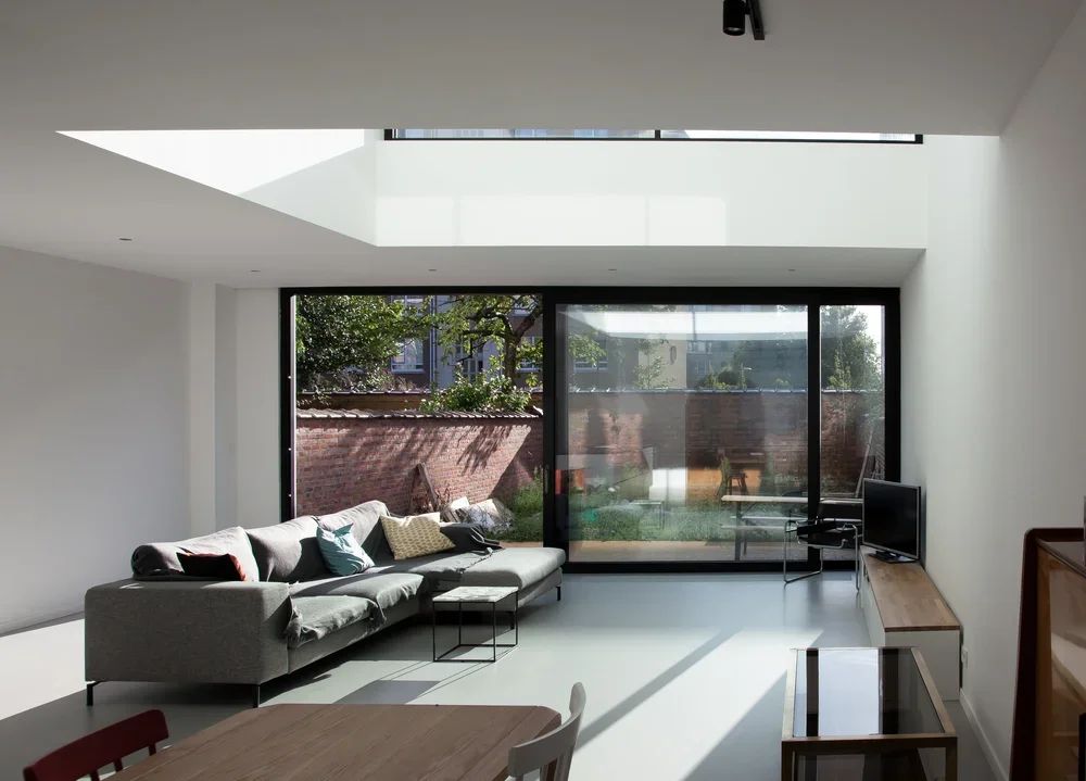 Modern living room with large glass sliding doors, a gray sofa with assorted pillows, a wooden TV stand with a small TV, and a dining table with chairs, bathed in natural sunlight.