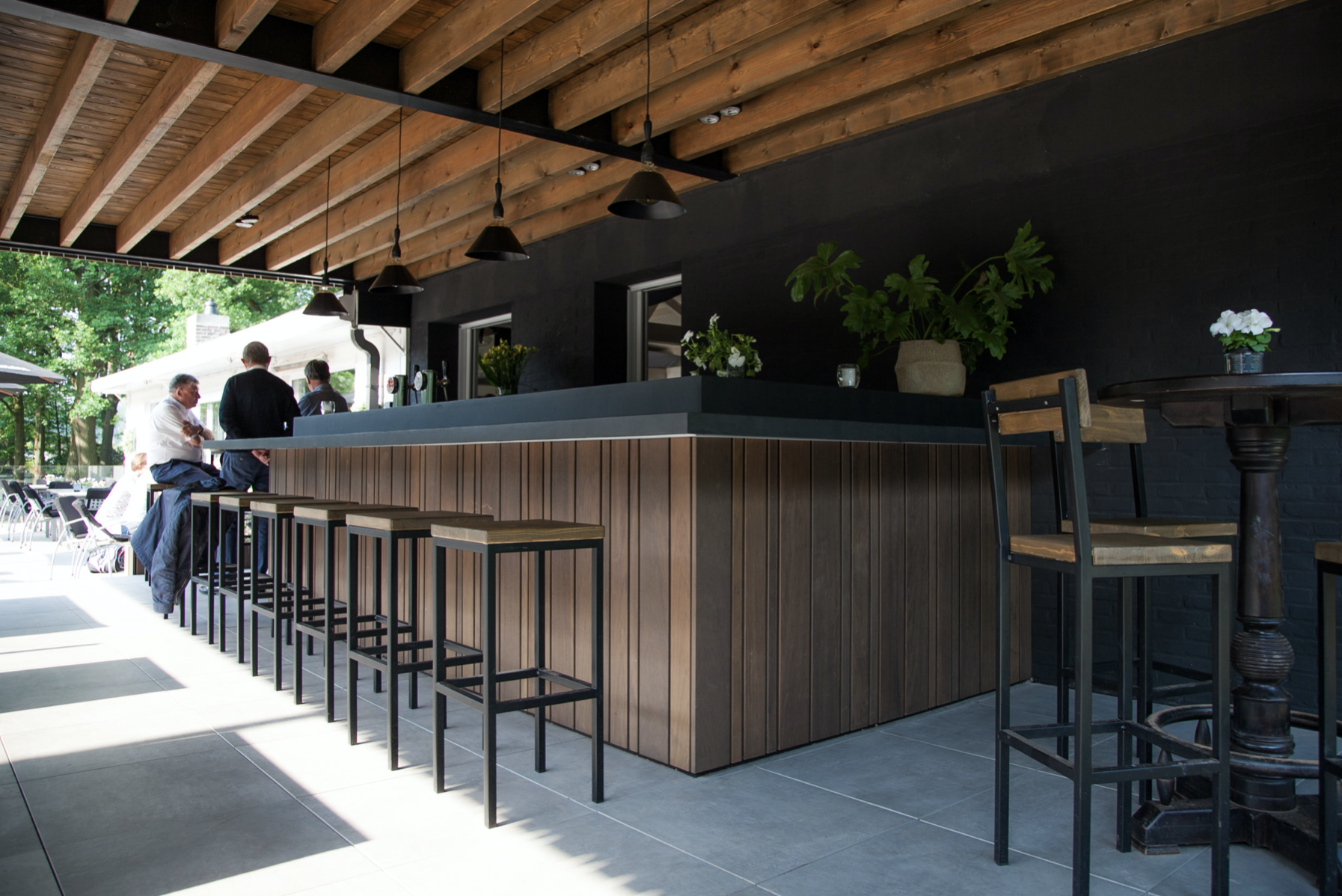 Modern bar and lounge interior design with a custom wooden counter, high stools, and large windows, crafted by Tom Naeyaert Architecture in Antwerp.