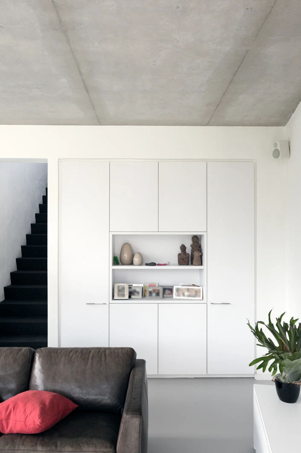 Modern living room with white built-in shelves, decorative stones and sculptures, a dark leather sofa with a red cushion, and a potted plant on a white side table. Stairs with black steps are visible on the left, and a speaker is mounted in the top right corner.