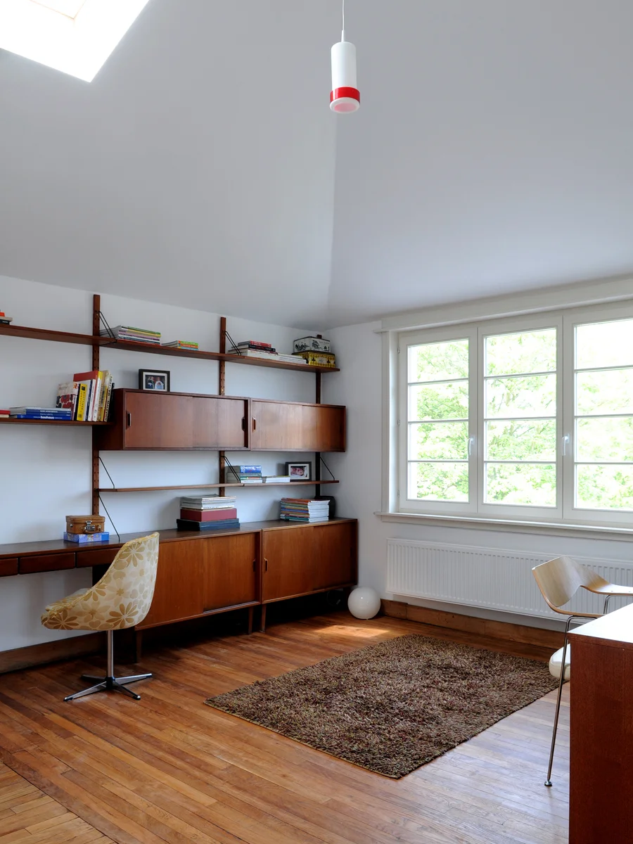 A bright room with a large window, wooden floor, a mid-century modern desk with a yellow floral chair, and wooden wall shelves filled with books and framed photos. There's a brown rug on the floor, a white spherical lamp, and a simple brown chair.