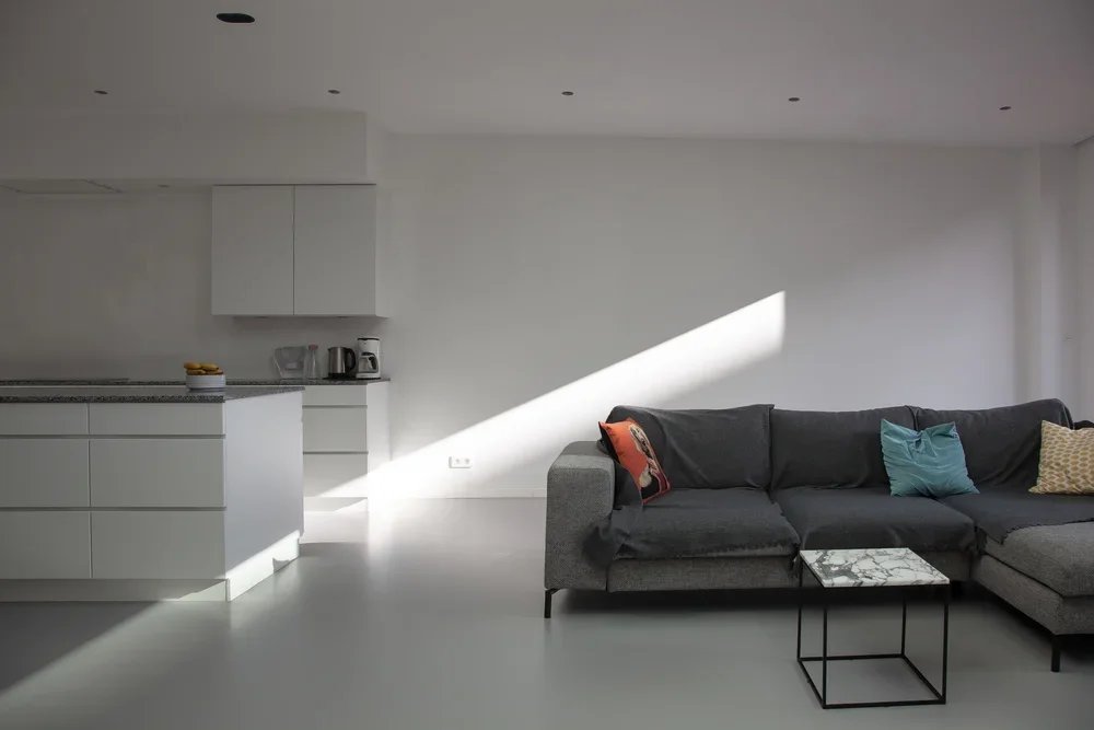 Minimalist living room with gray sectional sofa decorated with colorful pillows, marble coffee table, white kitchen with cabinets, countertop with appliances, and a large diagonal beam of sunlight on the wall.