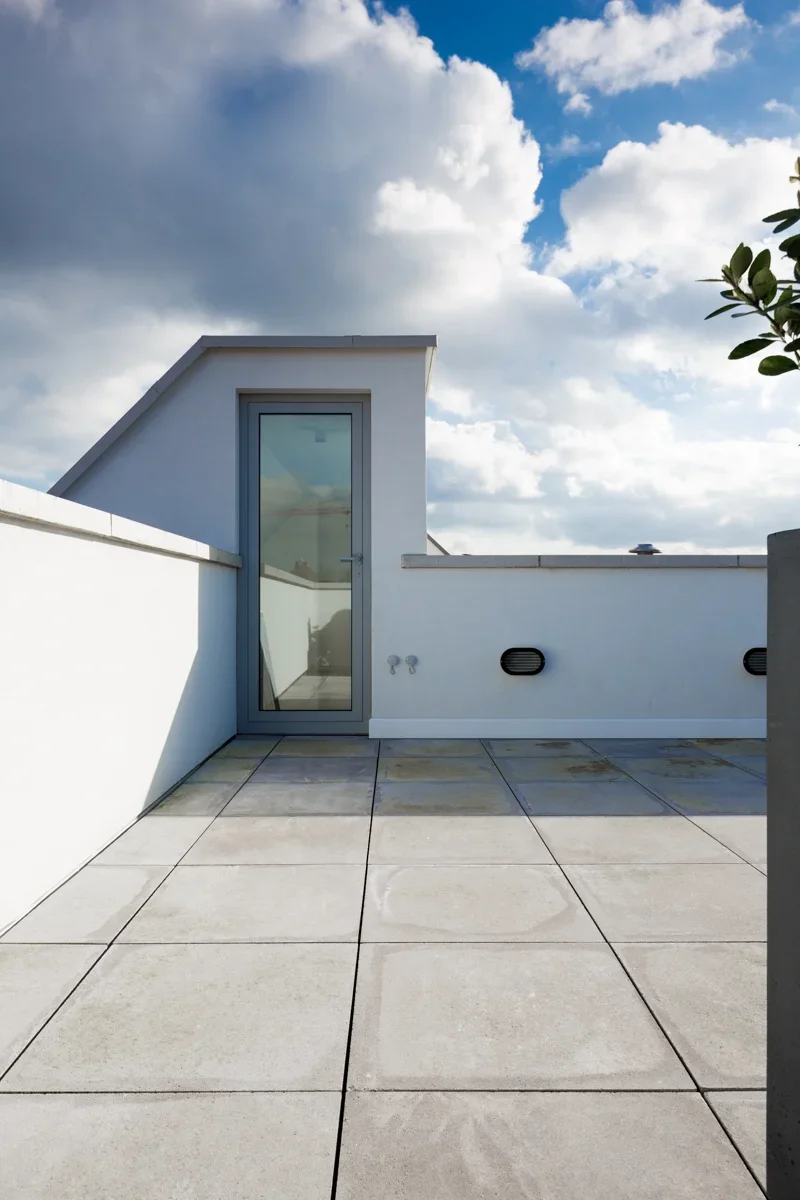 Empty rooftop terrace with large tiled floor, white walls, glass door, and partly cloudy sky.