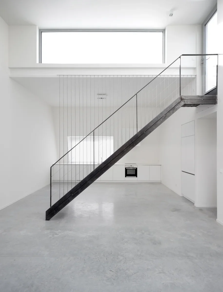 Empty minimalistic white room with concrete floor and a staircase with black railing and dark wooden steps leading up to a second level, large rectangular window at the top, and a kitchen area with white cabinets and a built-in oven.