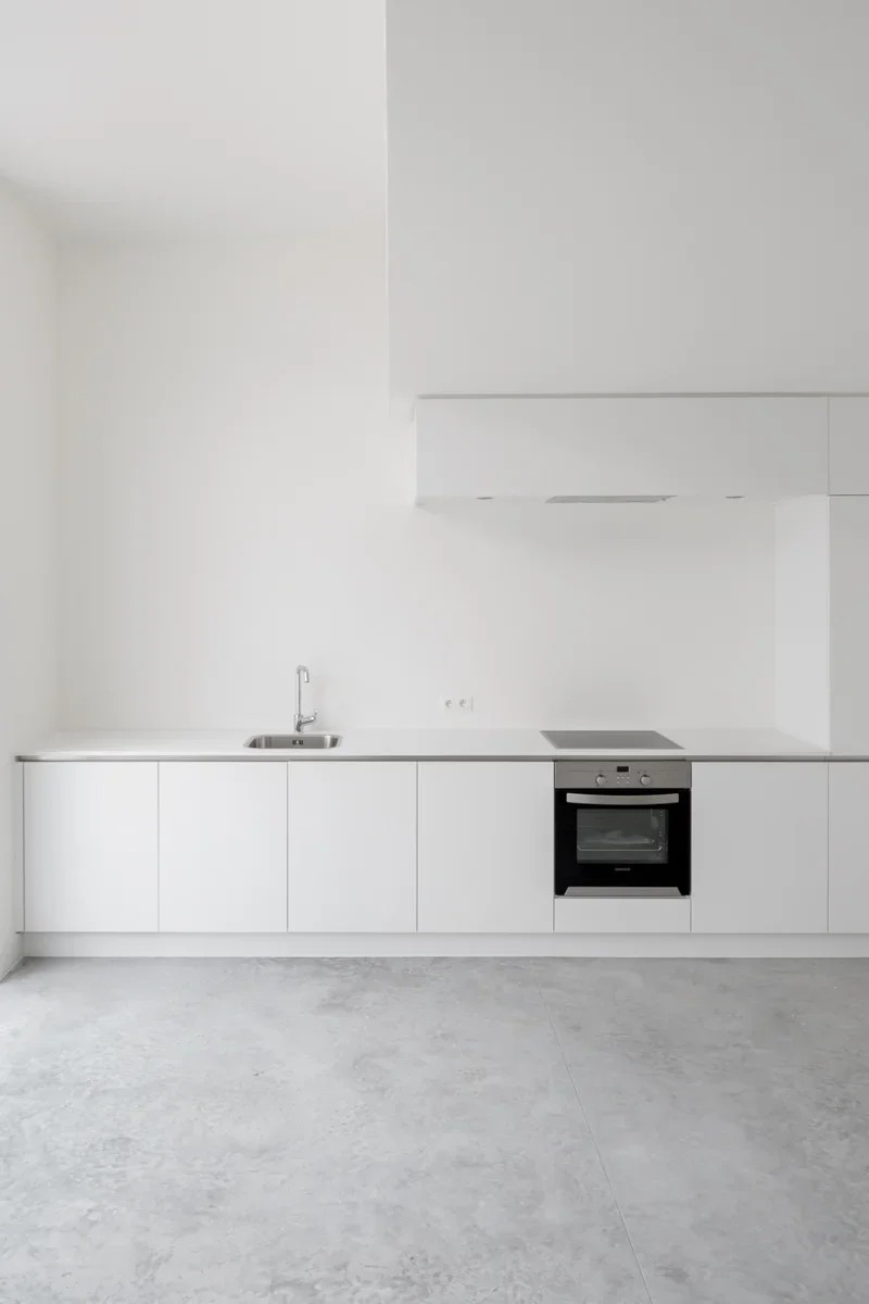 Minimalist white kitchen with built-in cabinets, a small sink, and an oven, with a light-colored floor and plain walls.