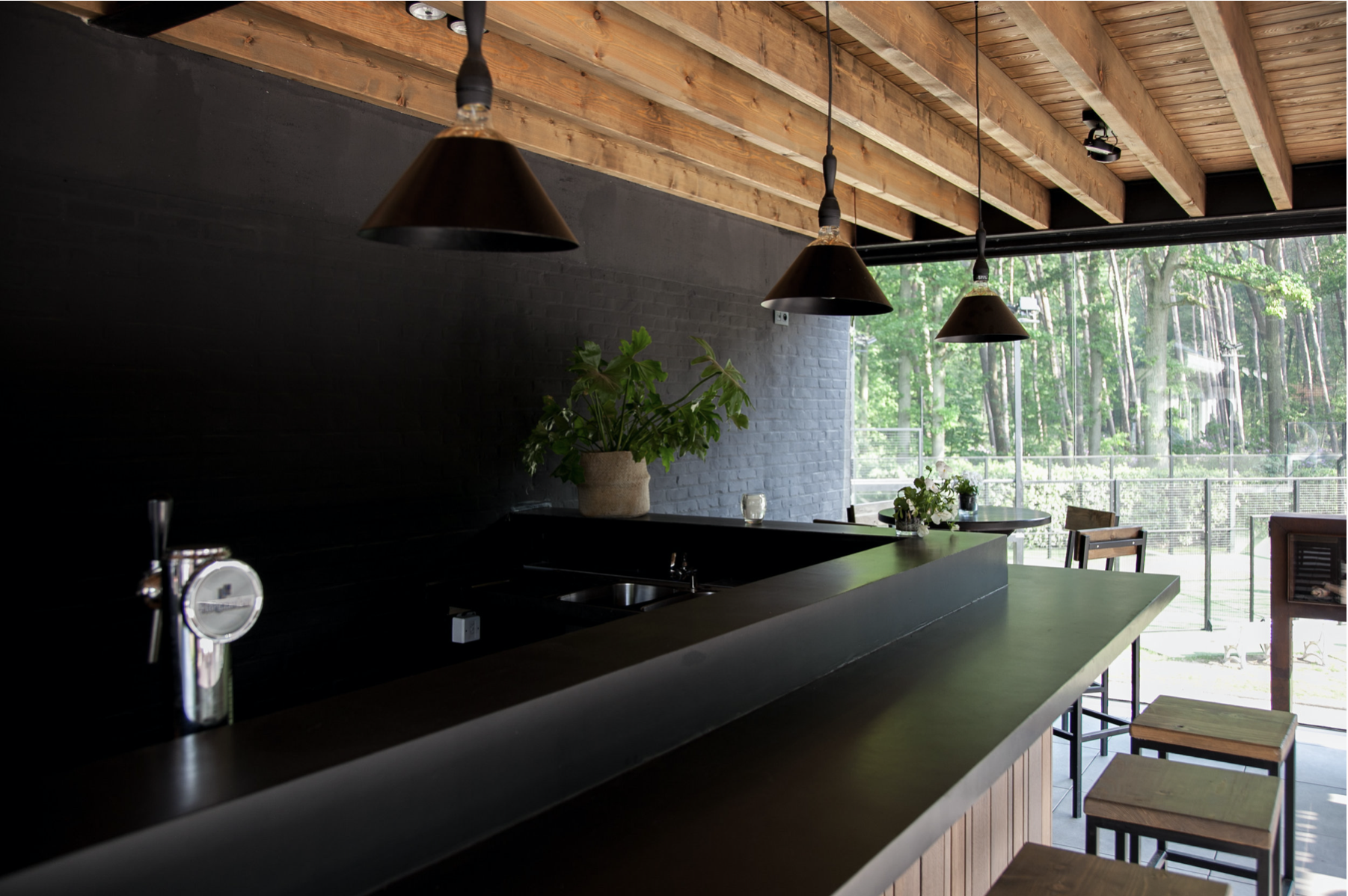 Contemporary clubhouse interior design featuring a long black counter, wooden ceiling beams, and industrial pendant lights, by Tom Naeyaert Architecture.