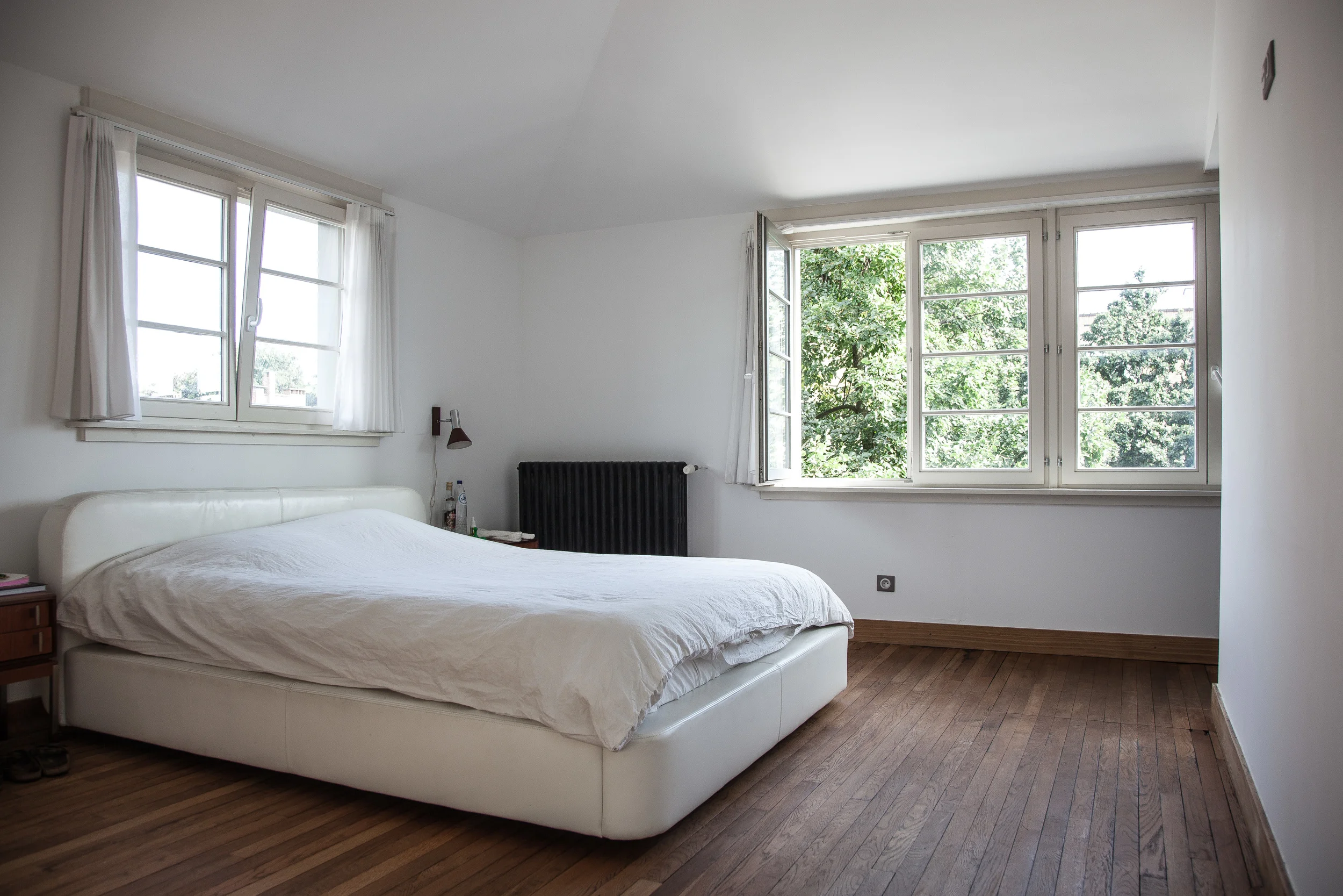 A minimalist bedroom with white walls, a large window with open white curtains, a white bed with a simple headboard, a small bedside table, a black radiator, wooden flooring, and view of green trees outside.