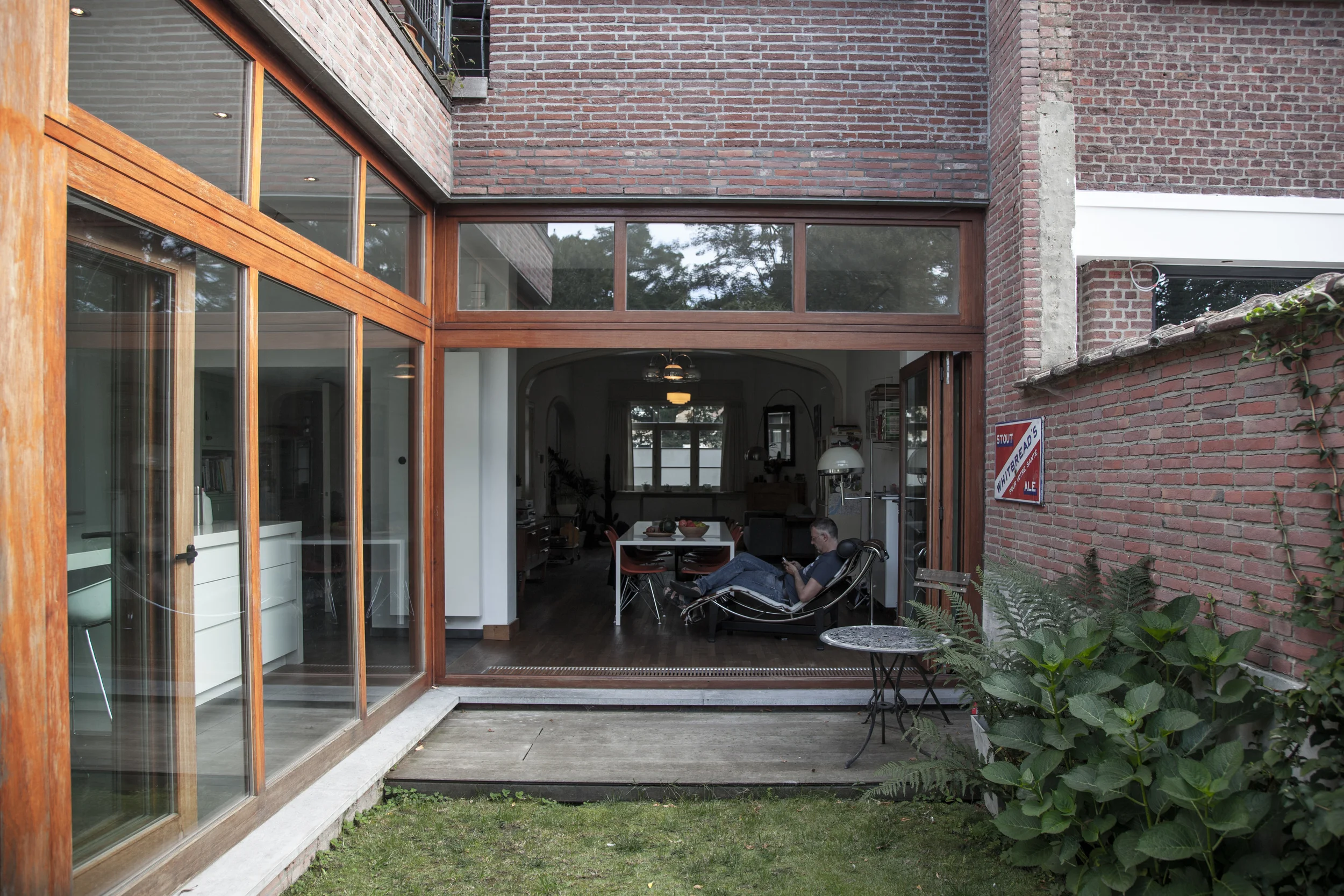 A modern home with large glass sliding doors opening into a living room, featuring brick and wooden exterior walls, with a small patio area outside and a man sitting on a lounge chair inside, looking at his phone, surrounded by plants.