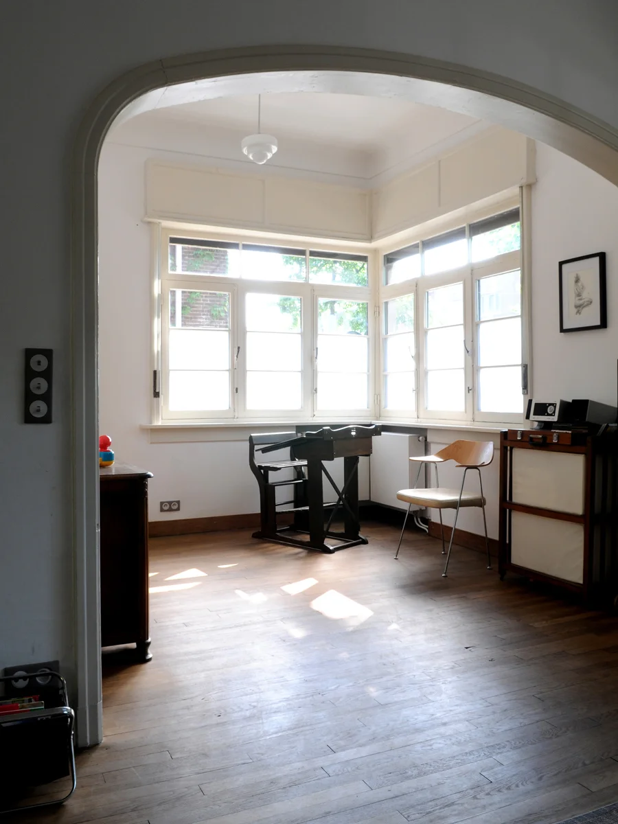Empty room with large bay window, wooden floor, and minimal furniture including a small table, a chair, and a speaker, illuminated by natural light.