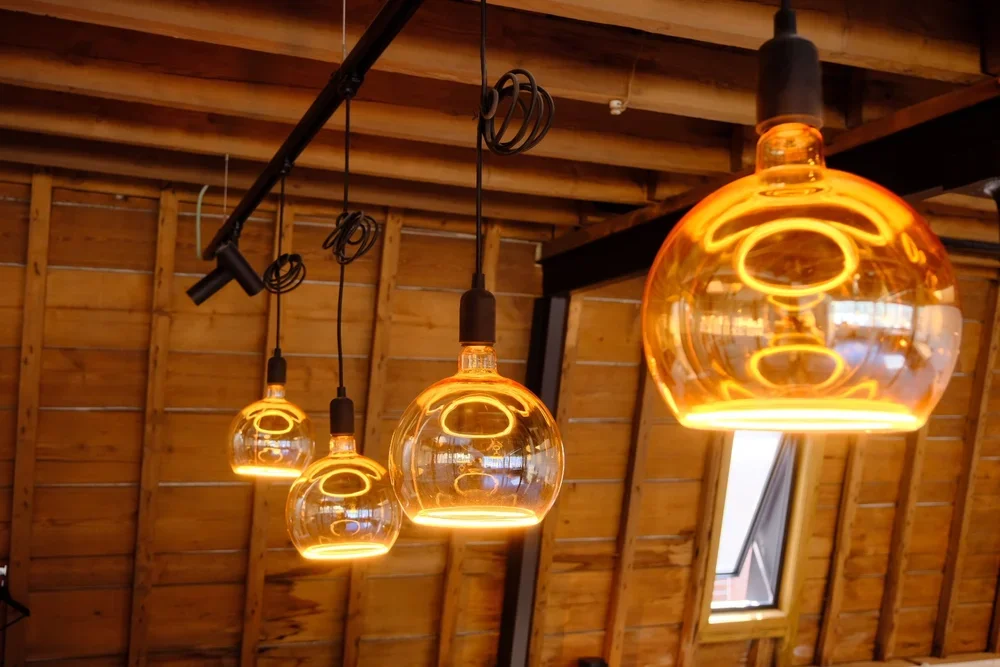 Bespoke amber-colored glass pendant lights hanging from a black track on a wooden ceiling in a modern rustic interior, by Tom Naeyaert Architecture.
