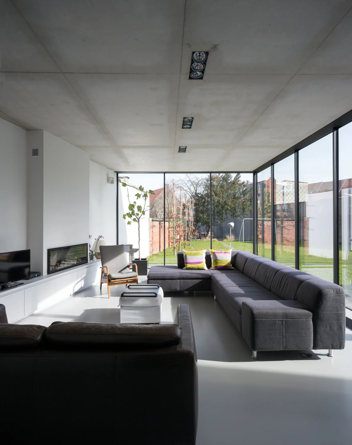 Modern living room with large glass windows, gray sectional sofa with colorful pillows, black leather sofa, white walls, minimalist fireplace, armchair, and outdoor garden with trees and grass visible through the windows.
