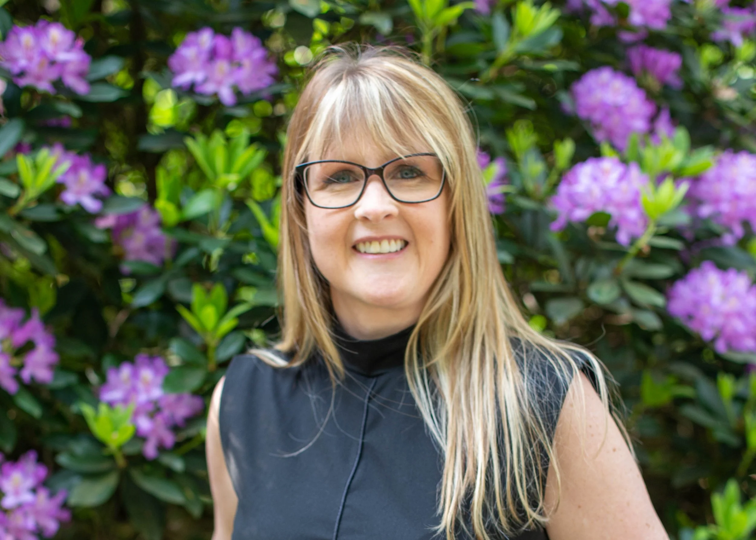 A woman with blonde hair and glasses smiling outdoors with purple flowers and green foliage in the background.