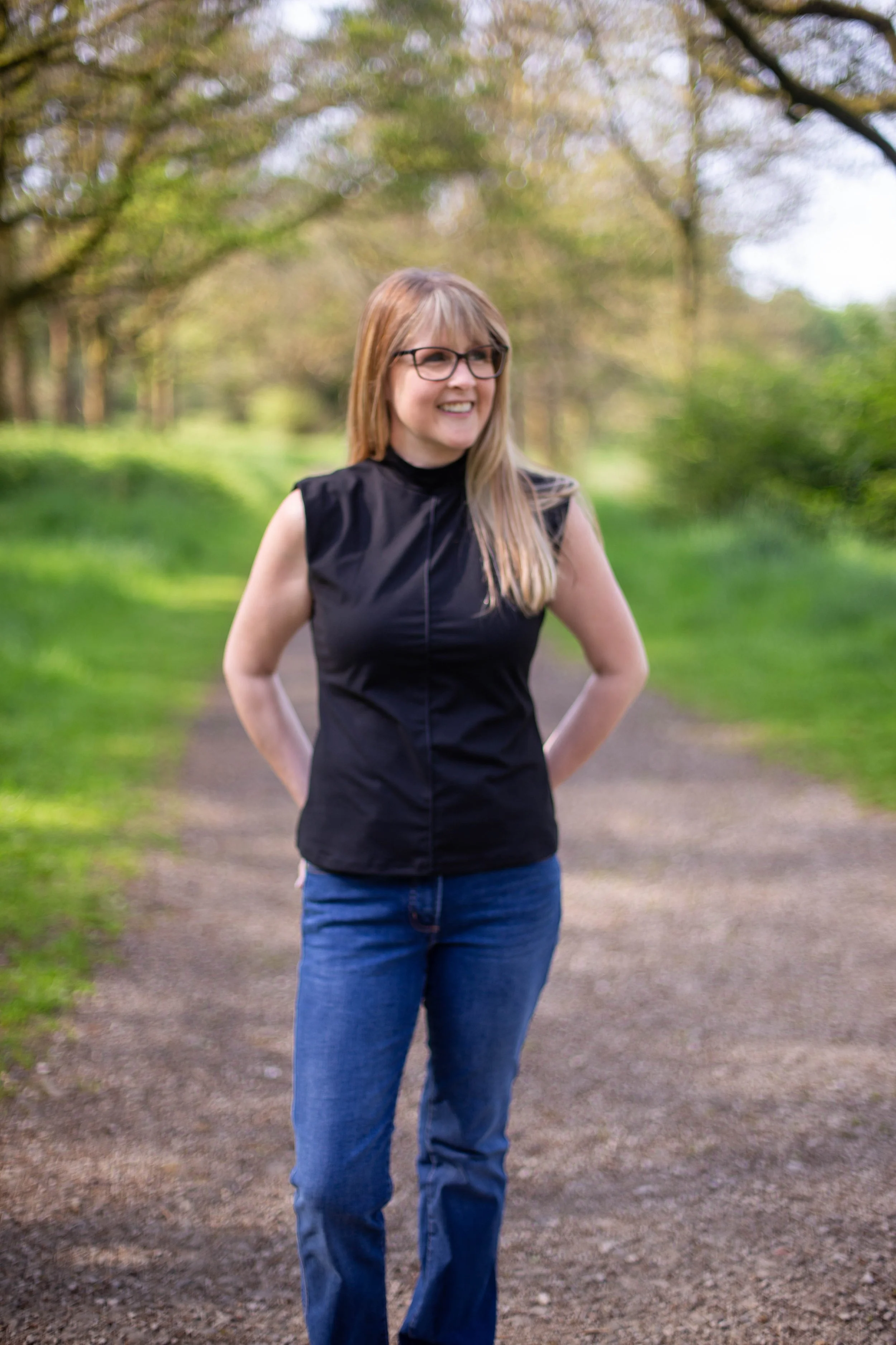 A woman with long blonde hair, glasses, wearing a black sleeveless top and blue jeans, smiling and standing on a dirt path in a park with green grass and trees in the background.