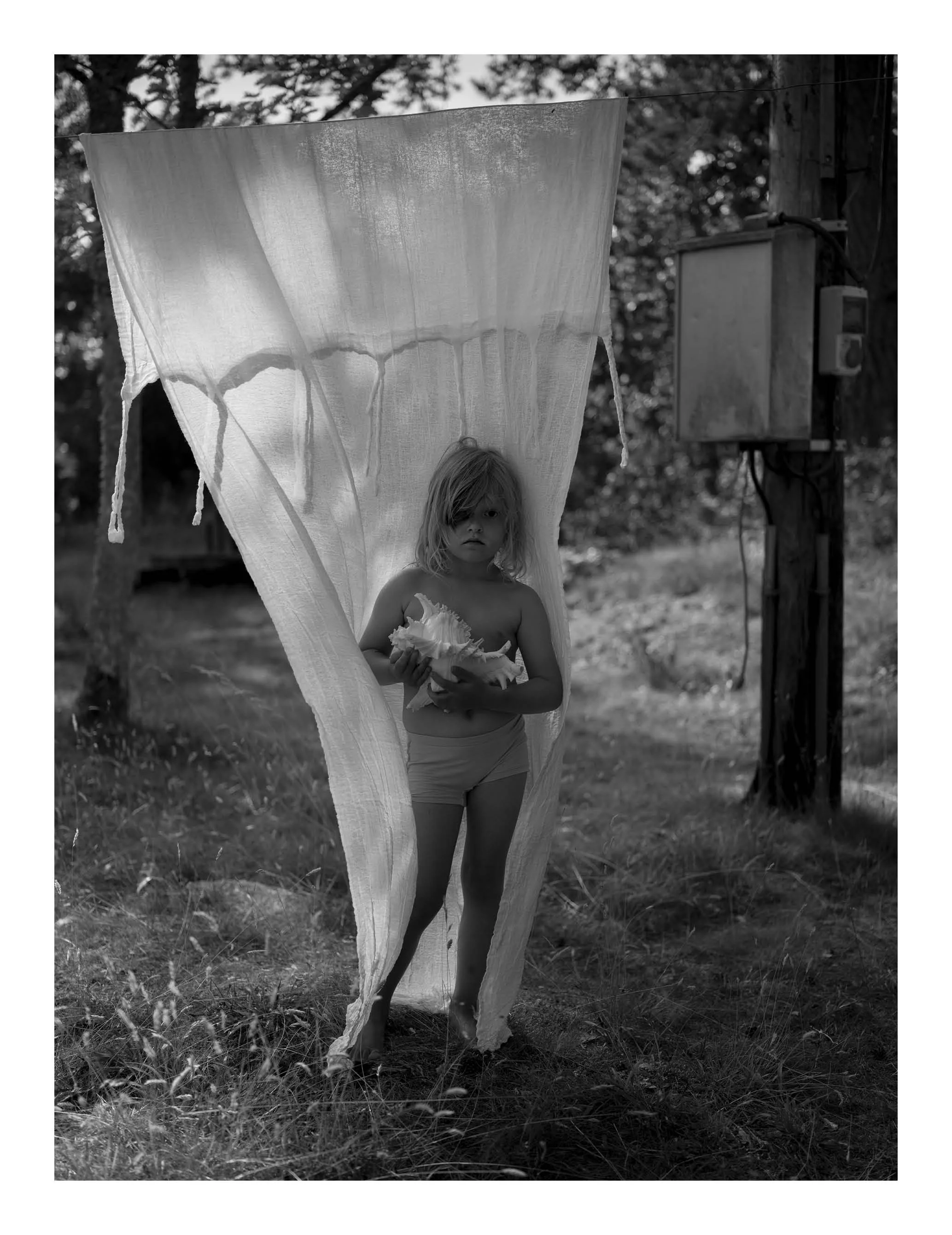 A young girl with long hair standing outdoors behind a large piece of fabric, holding an object in her hands, with trees and a utility box in the background, in black and white.