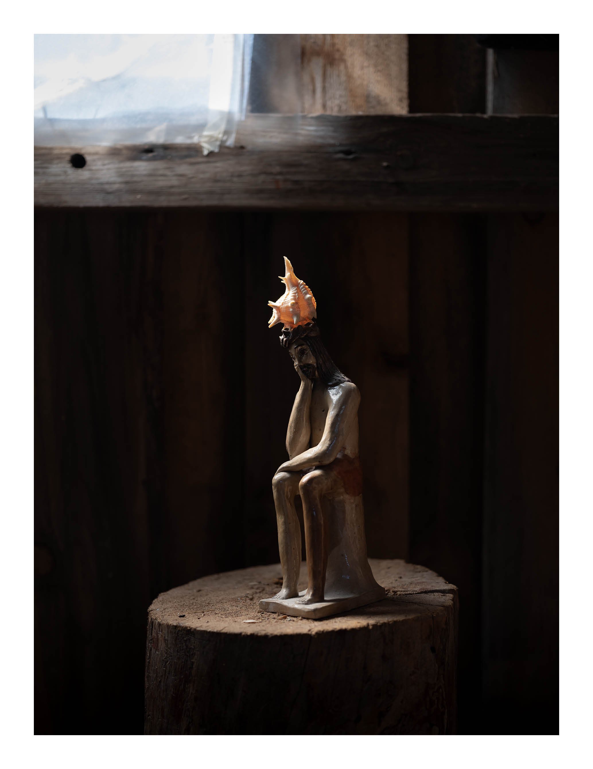 Wooden sculpture of a woman sitting with her head resting on her hand, holding a conch shell on top of her head, placed on a wooden base and displayed on a tree stump.