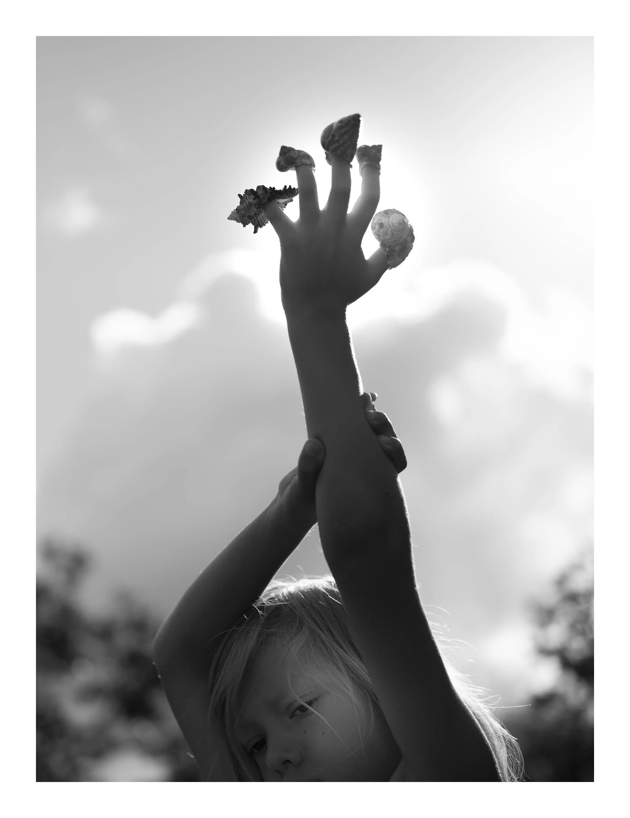 A young girl with blonde hair playfully shielding her face with her arm and lifting her hand, with sunflower petal-shaped rings on her fingers, standing outdoors under sunlight.