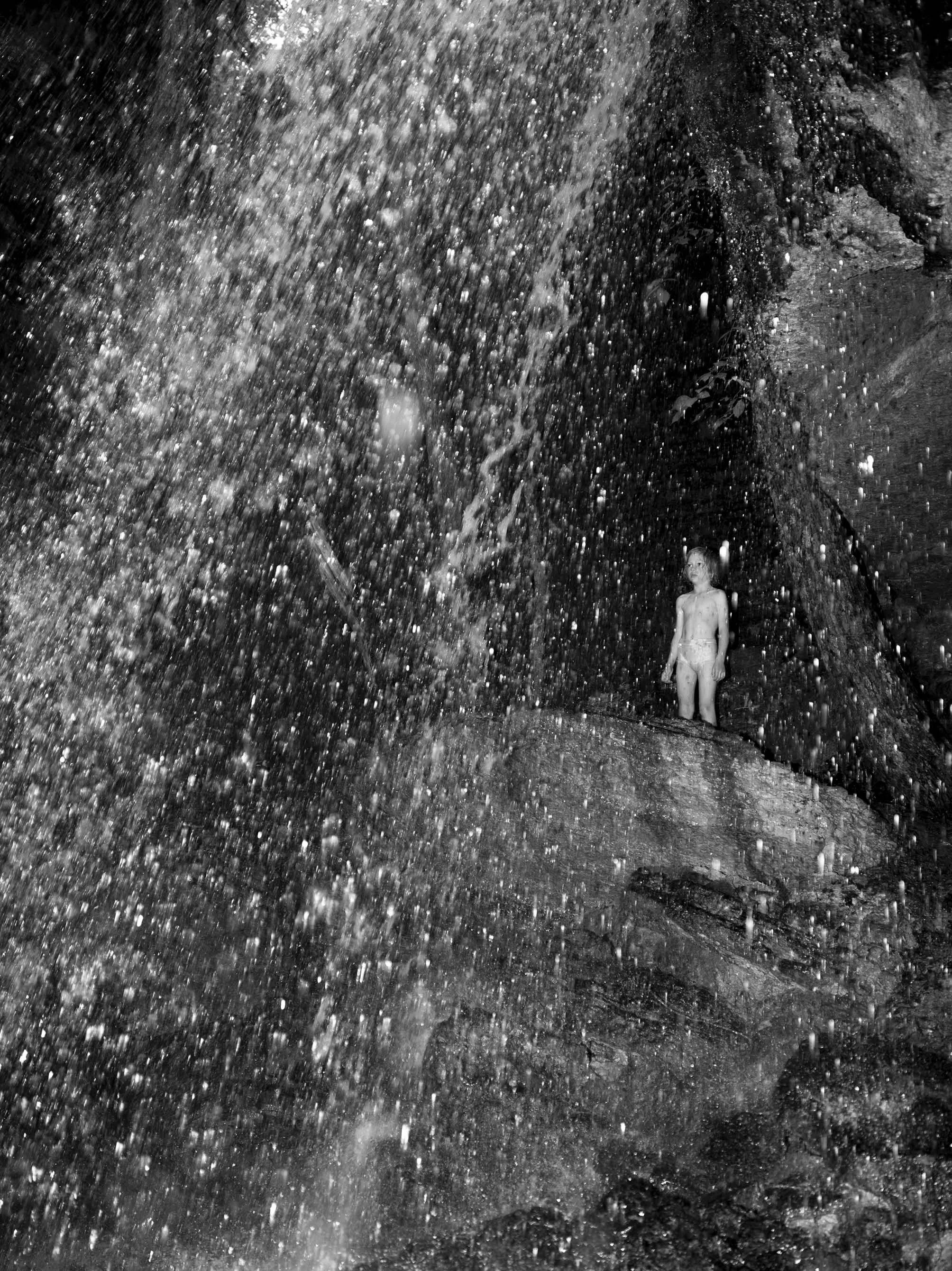 A young boy standing underneath a waterfall of water, with rocks and plants around him, in a natural outdoor setting.