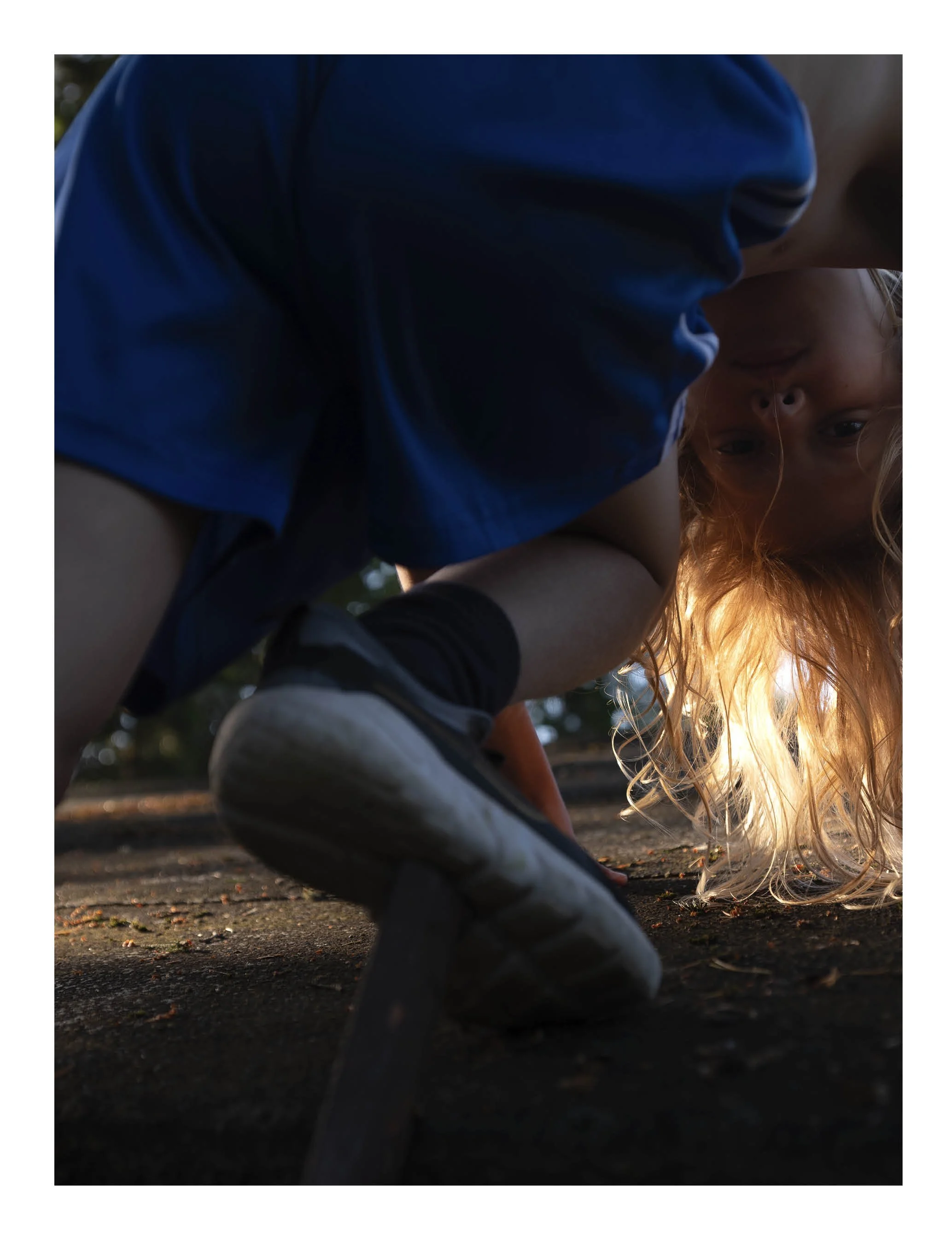A young girl with long blond hair is crouching on the ground outdoors, looking down. She is wearing a light-colored shirt and has a gentle smile. The photo is taken from a low angle with sunlight illuminating her hair.