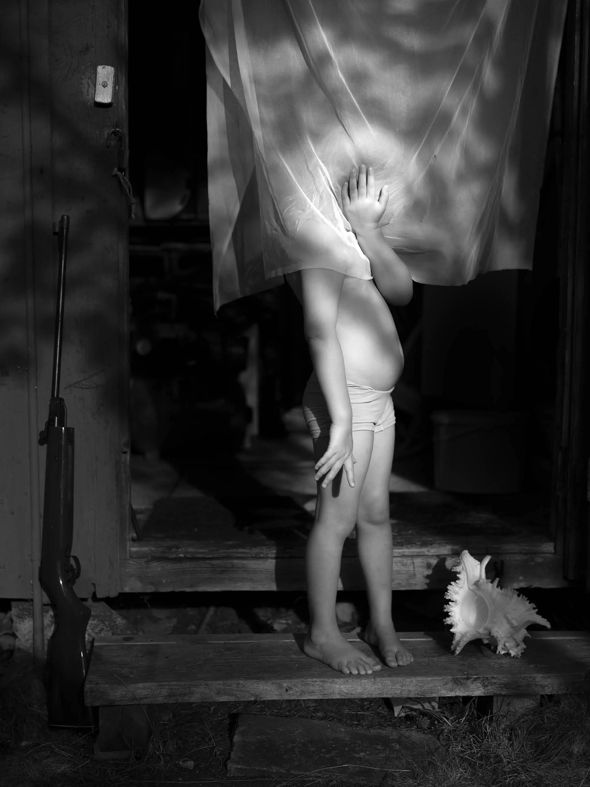 A black and white photo of a child with their face obscured, standing on a wooden platform in front of a curtain. The child's right hand is on the curtain and a large shell is on the ground nearby. There is a saw or similar tool leaning against the w